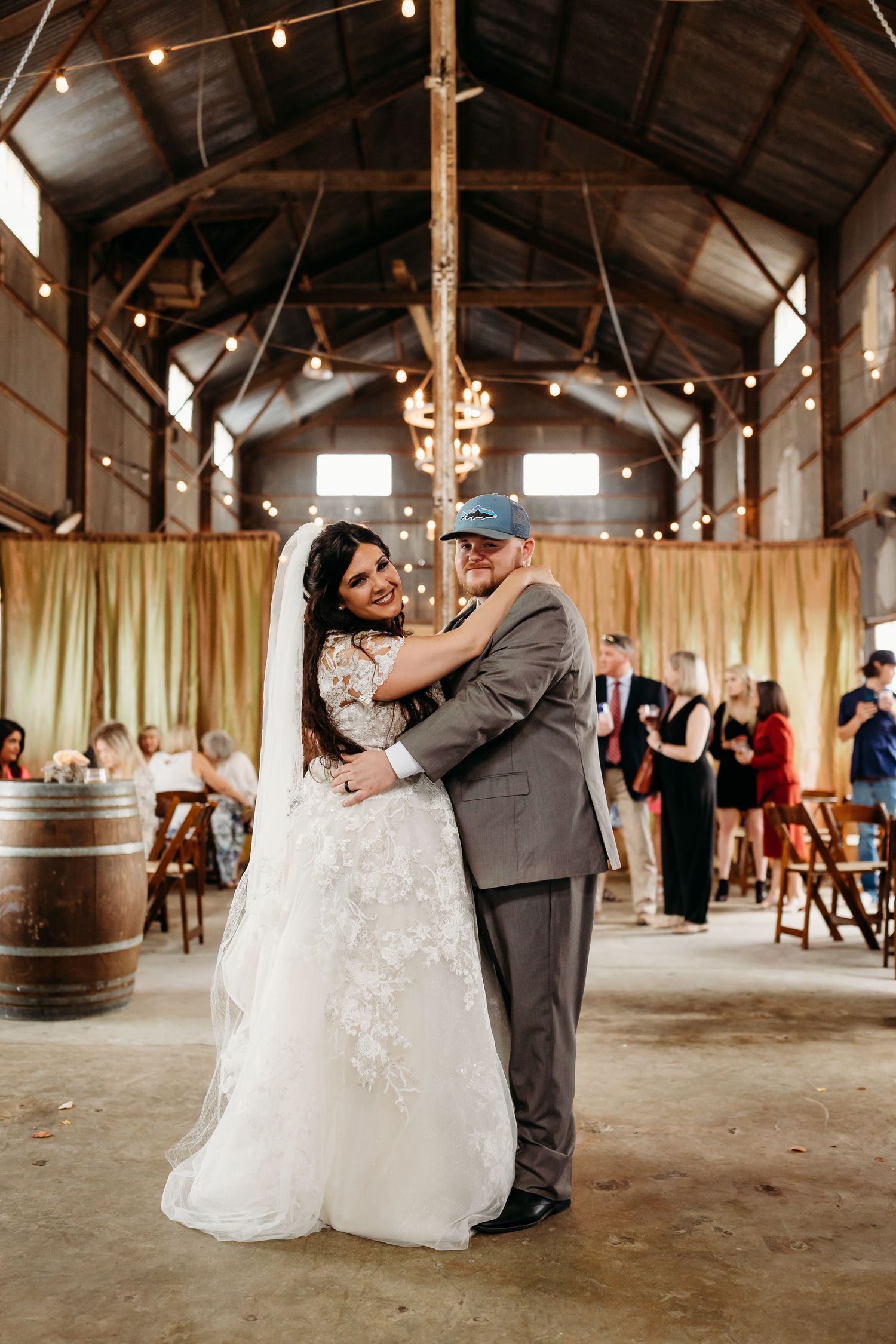 Couple shares an intimate dance moment in a barn with string lights and wooden barrels.