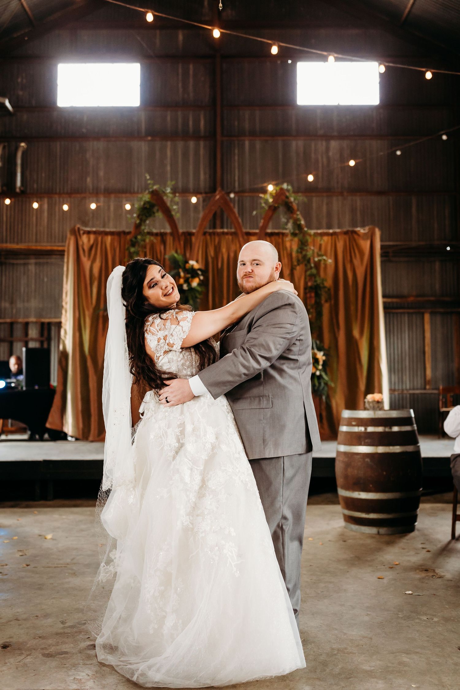 Dancing couple in formal attire inside a rustic barn venue with warm lighting and barrel decor.