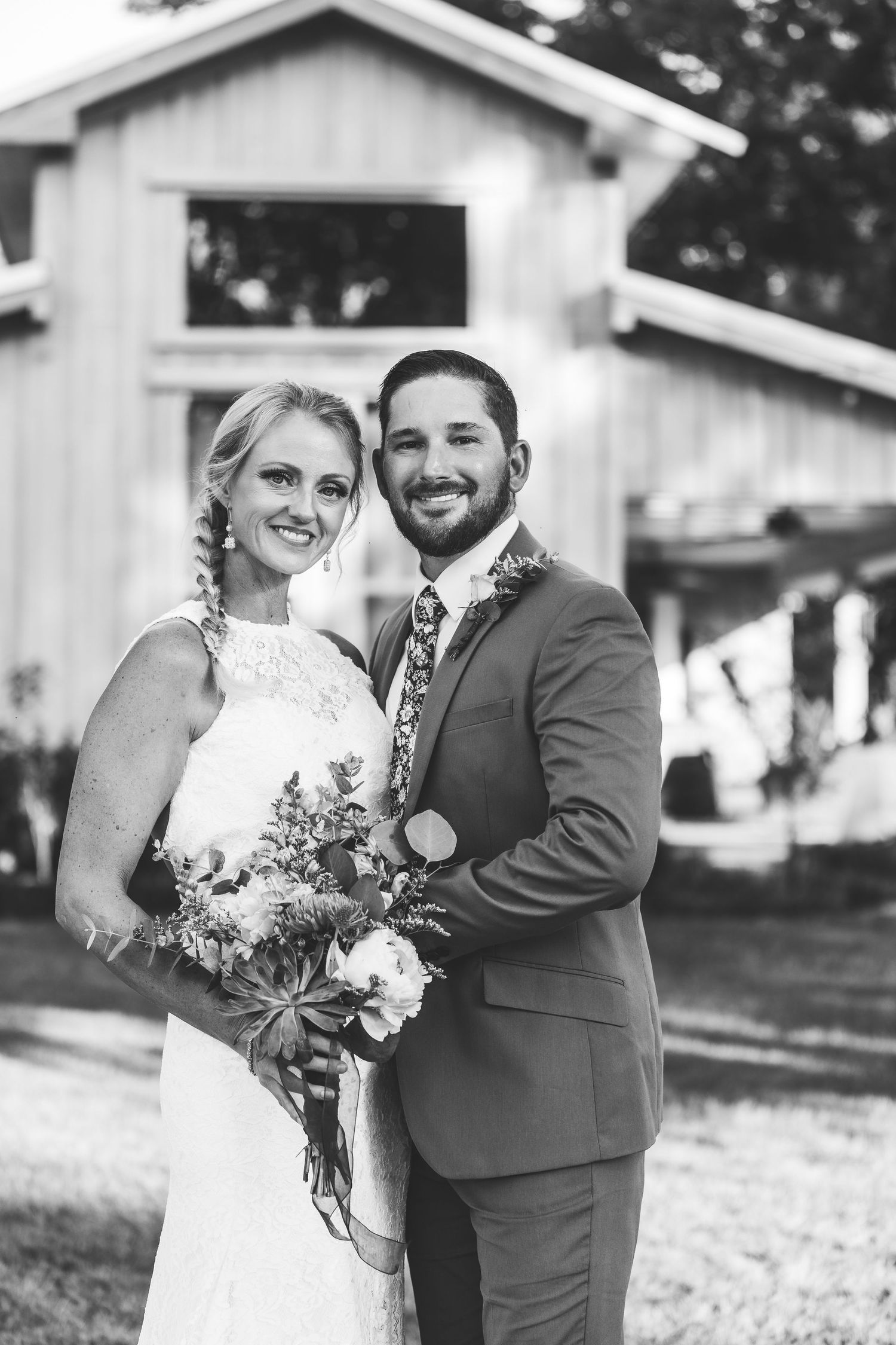 A wedding portrait in front of a white barn with a bouquet of mixed flowers and greenery.