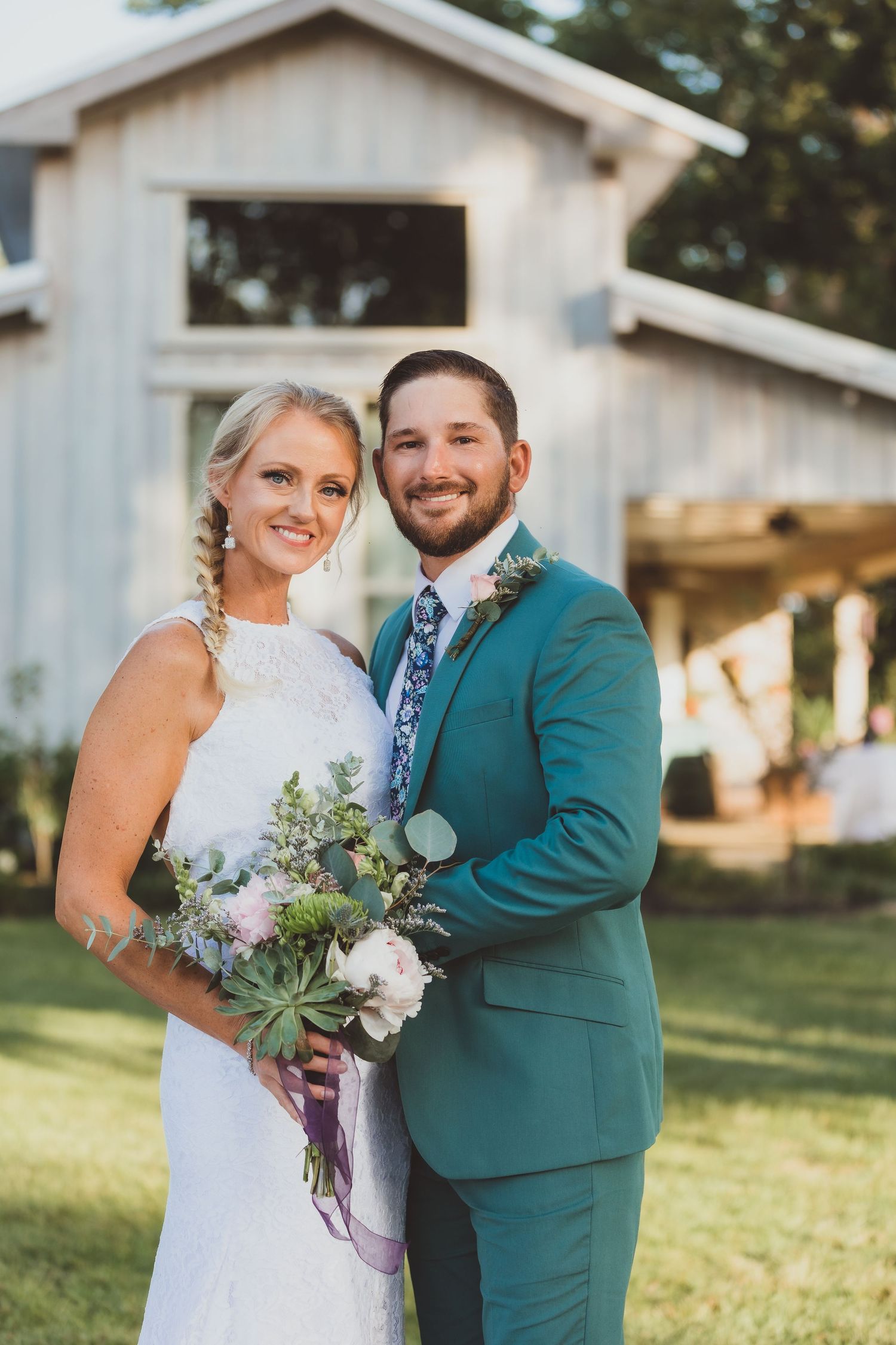 A newlywed couple stands in front of a rustic barn with the groom in a teal suit and bride holding a floral bouquet.