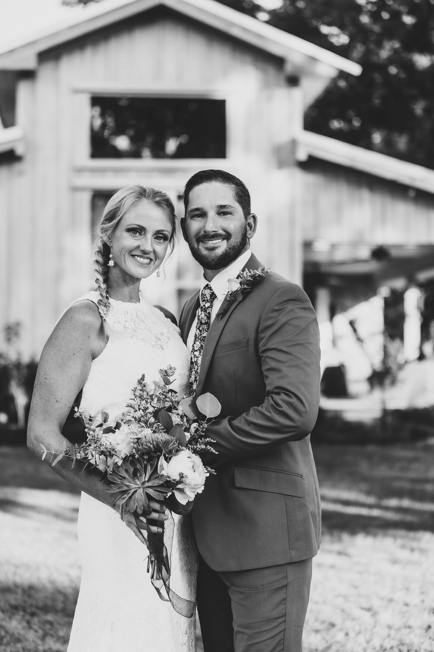 A black and white wedding portrait captures a couple smiling in front of a rustic barn setting.