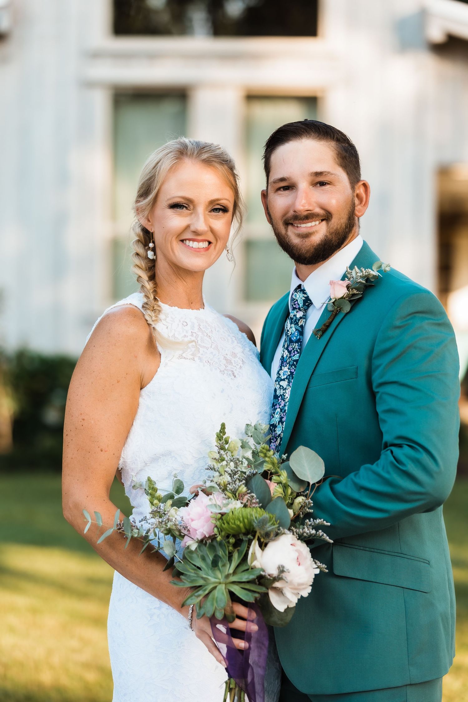 A wedding couple poses outdoors in elegant attire featuring a teal suit and white lace dress with a rustic bouquet.
