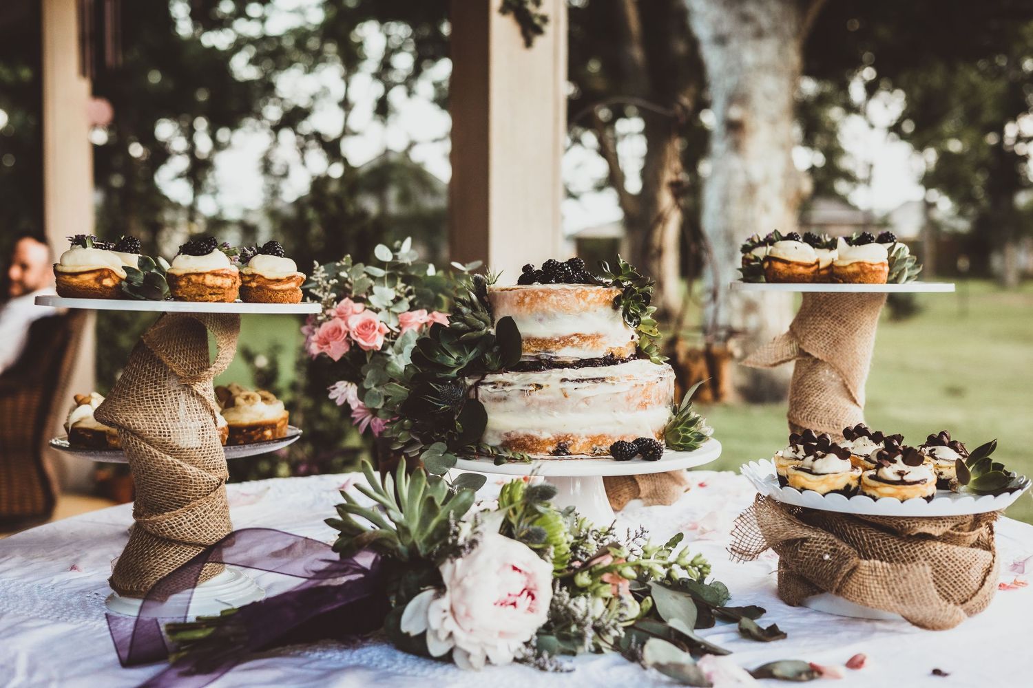 A rustic wedding dessert table displays tiered cakes and treats on wooden stands with floral decorations.