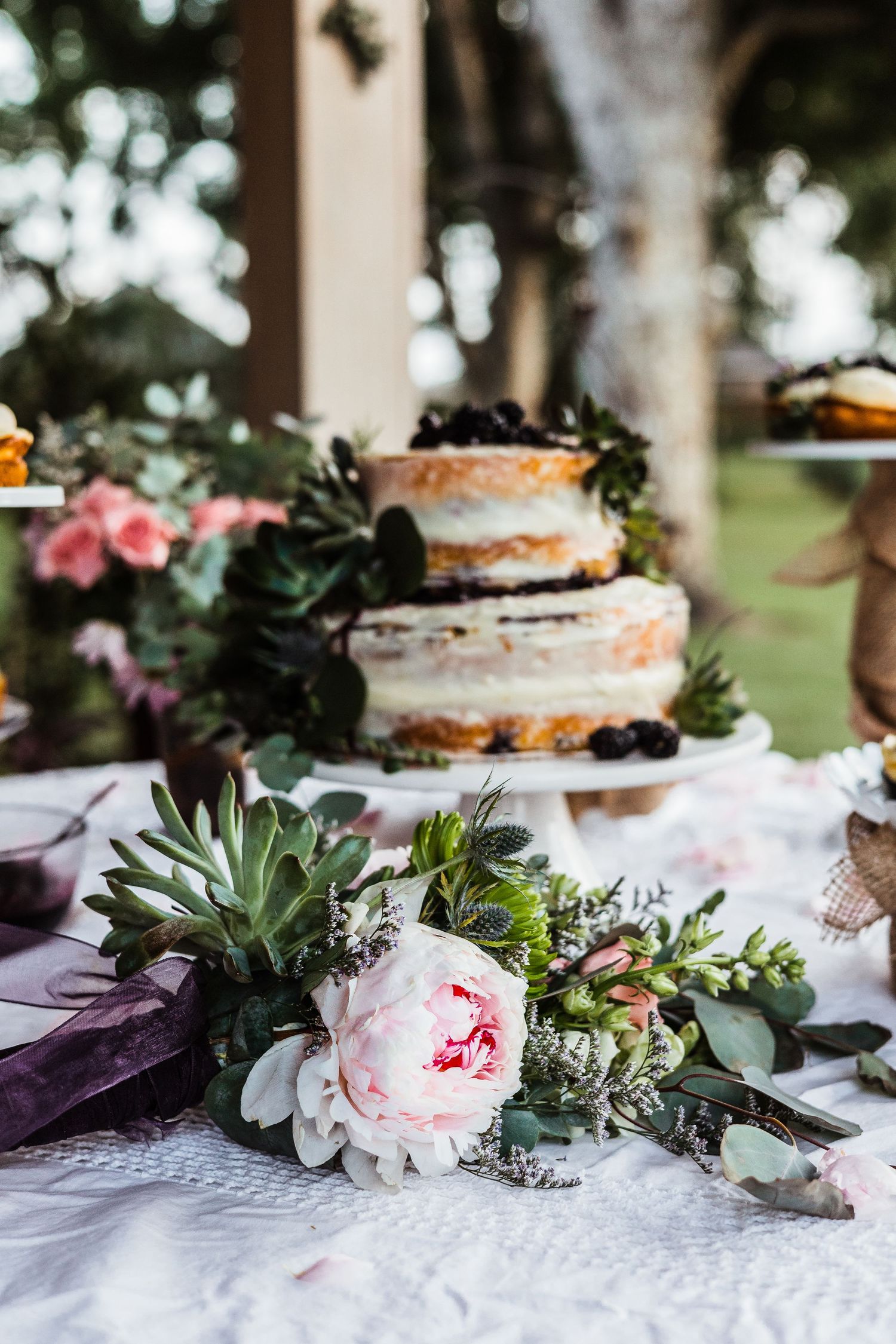 Semi-naked wedding cake adorned with succulents and pink peonies on a white tablecloth.