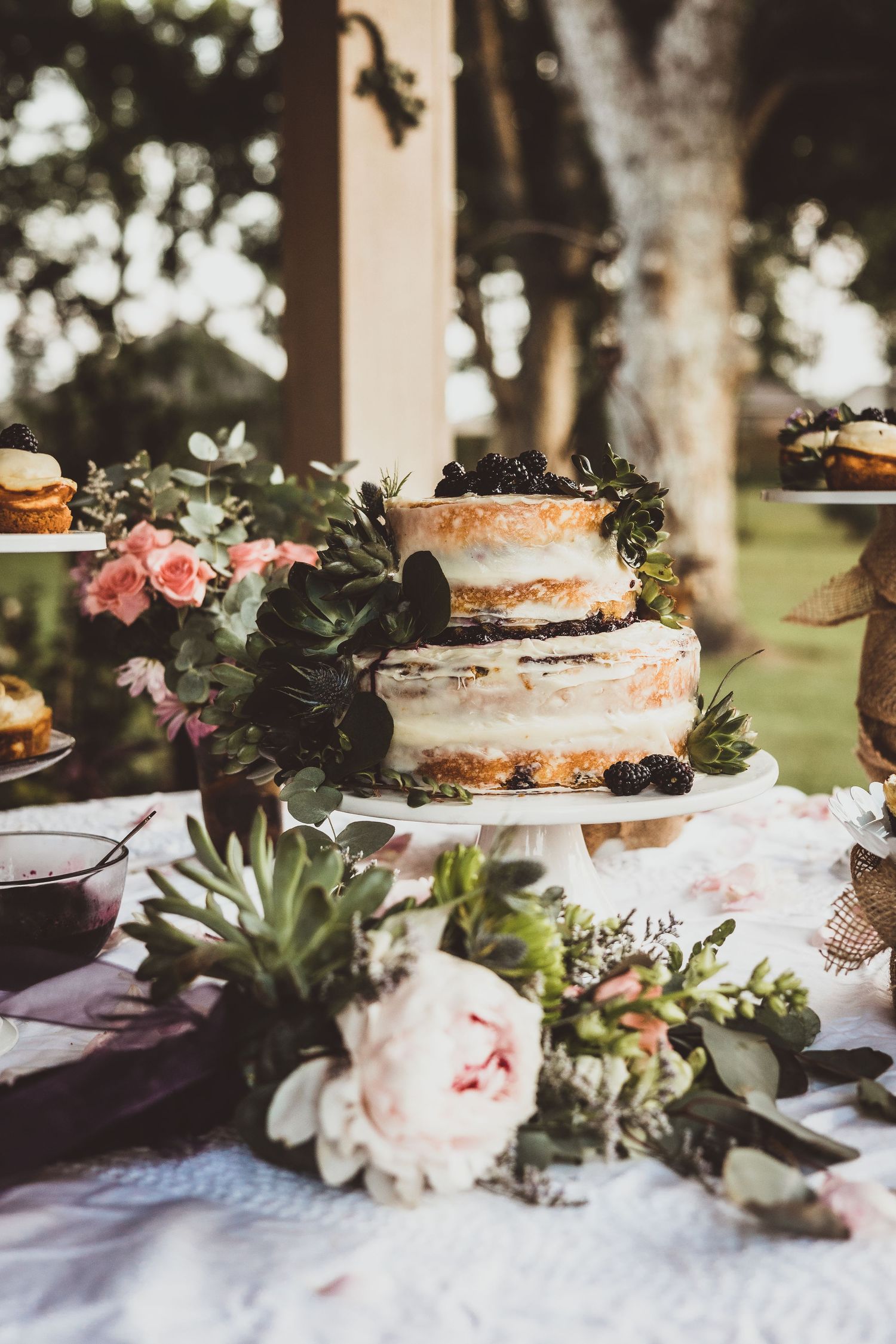 Rustic naked wedding cake decorated with fresh flowers and greenery on an outdoor dessert table.