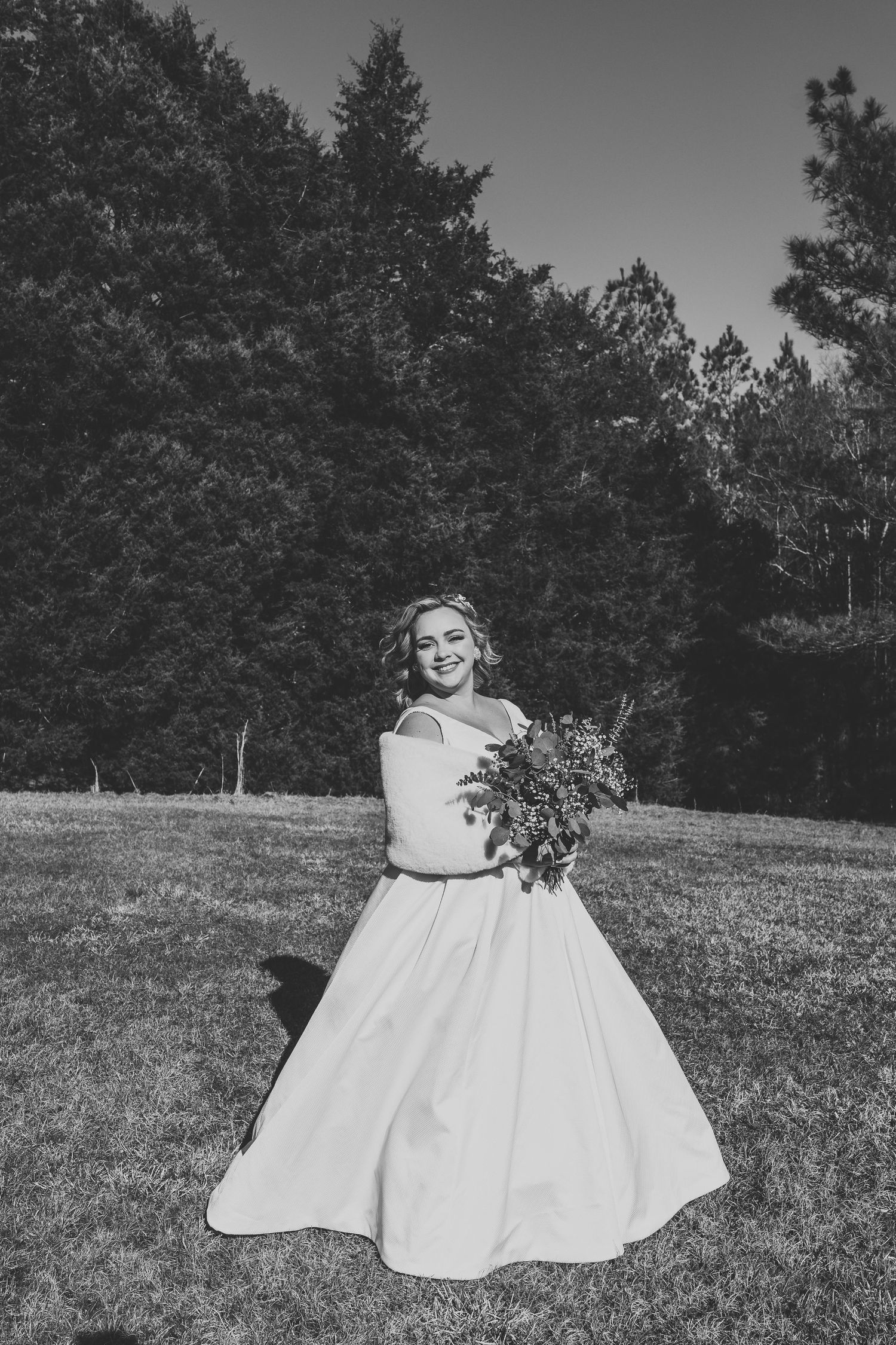A person in a white dress sitting on a grassy hillside surrounded by trees in black and white photography.