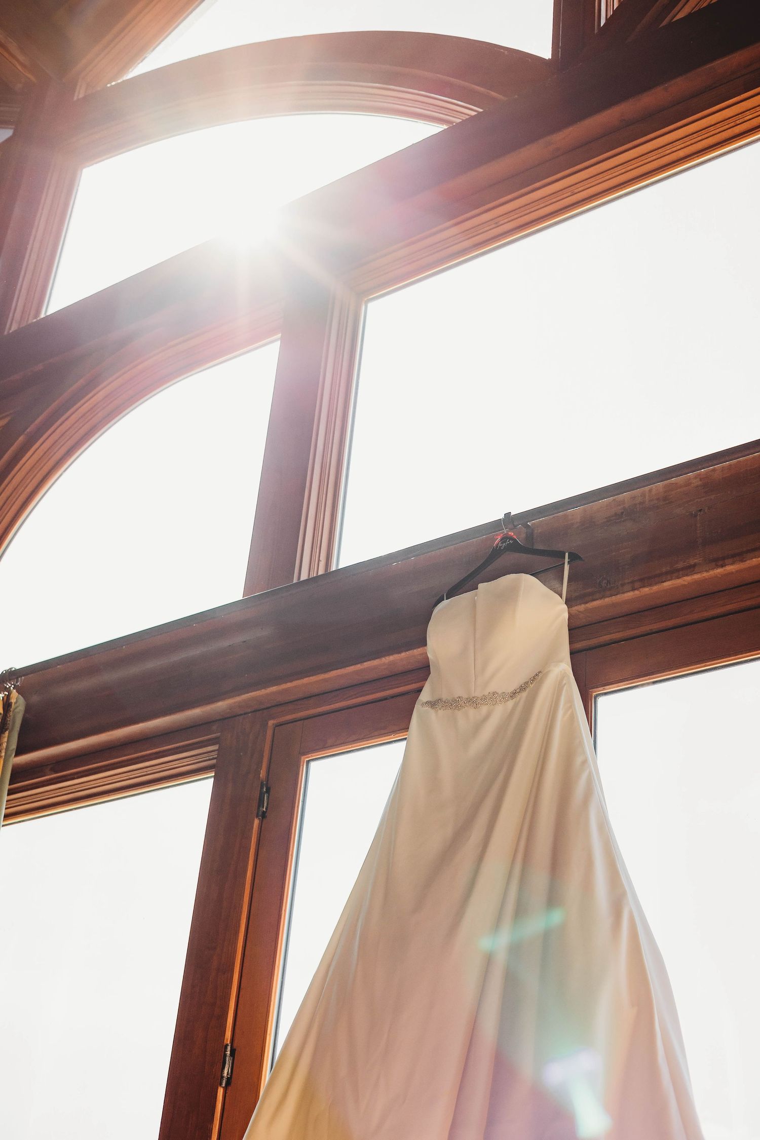 Wedding dress hanging in front of sunlit windows with wooden trim.