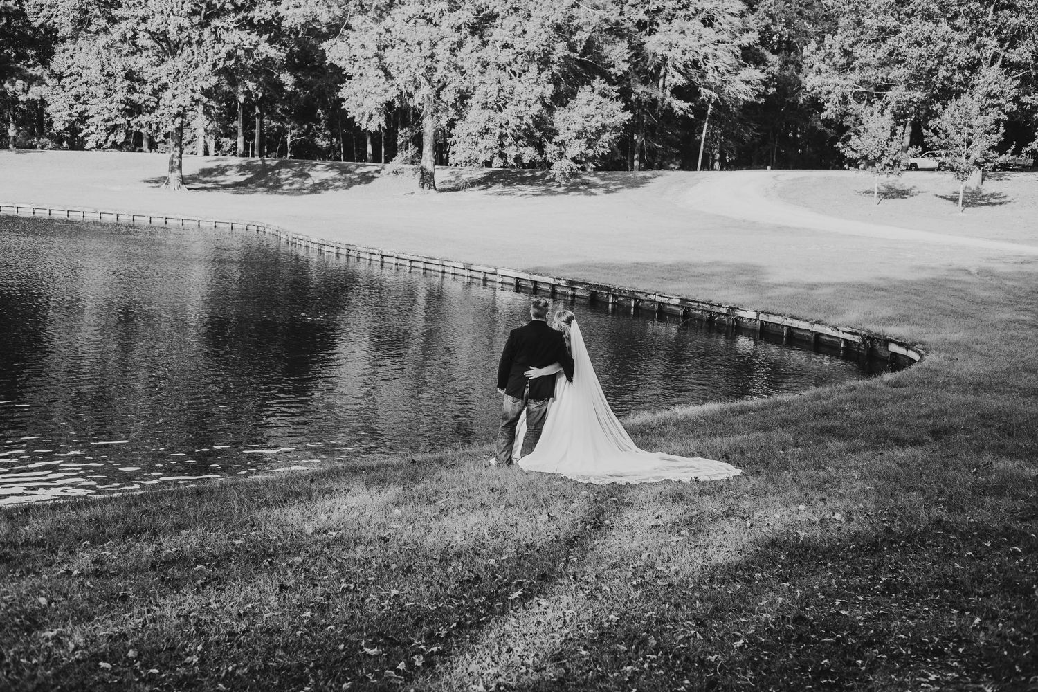 Black and white photo of newlyweds walking near a lake with trees in the background.