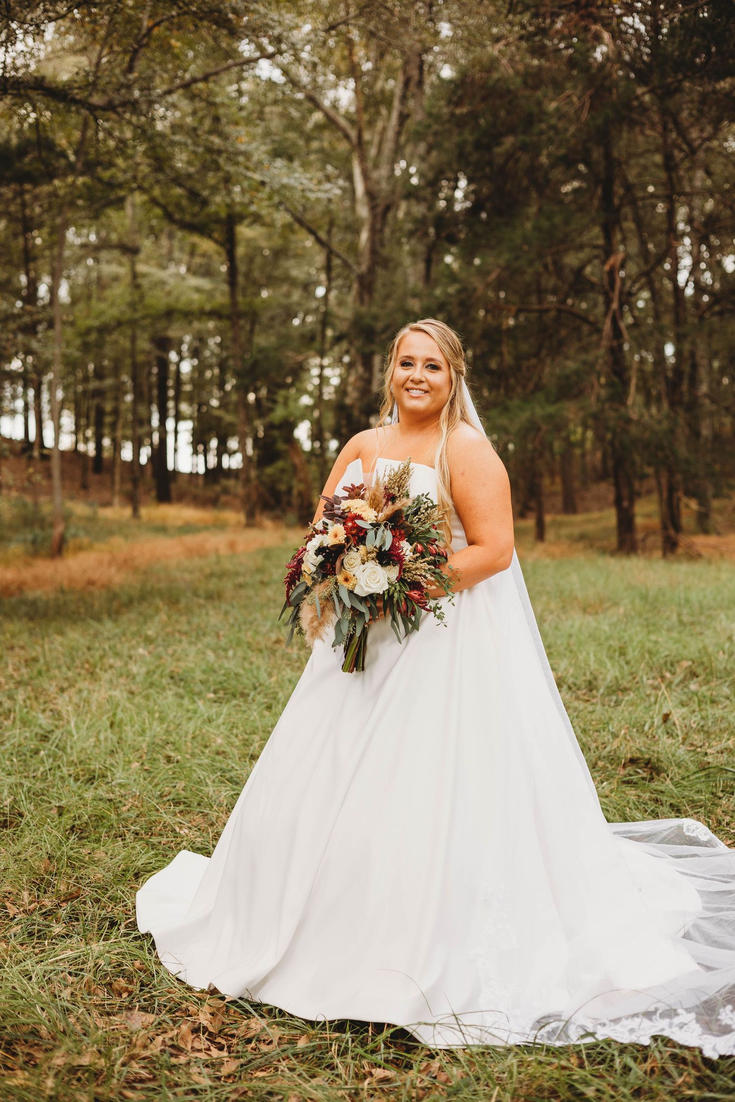 Bride in flowing white dress poses with autumn bouquet on grassy hillside.