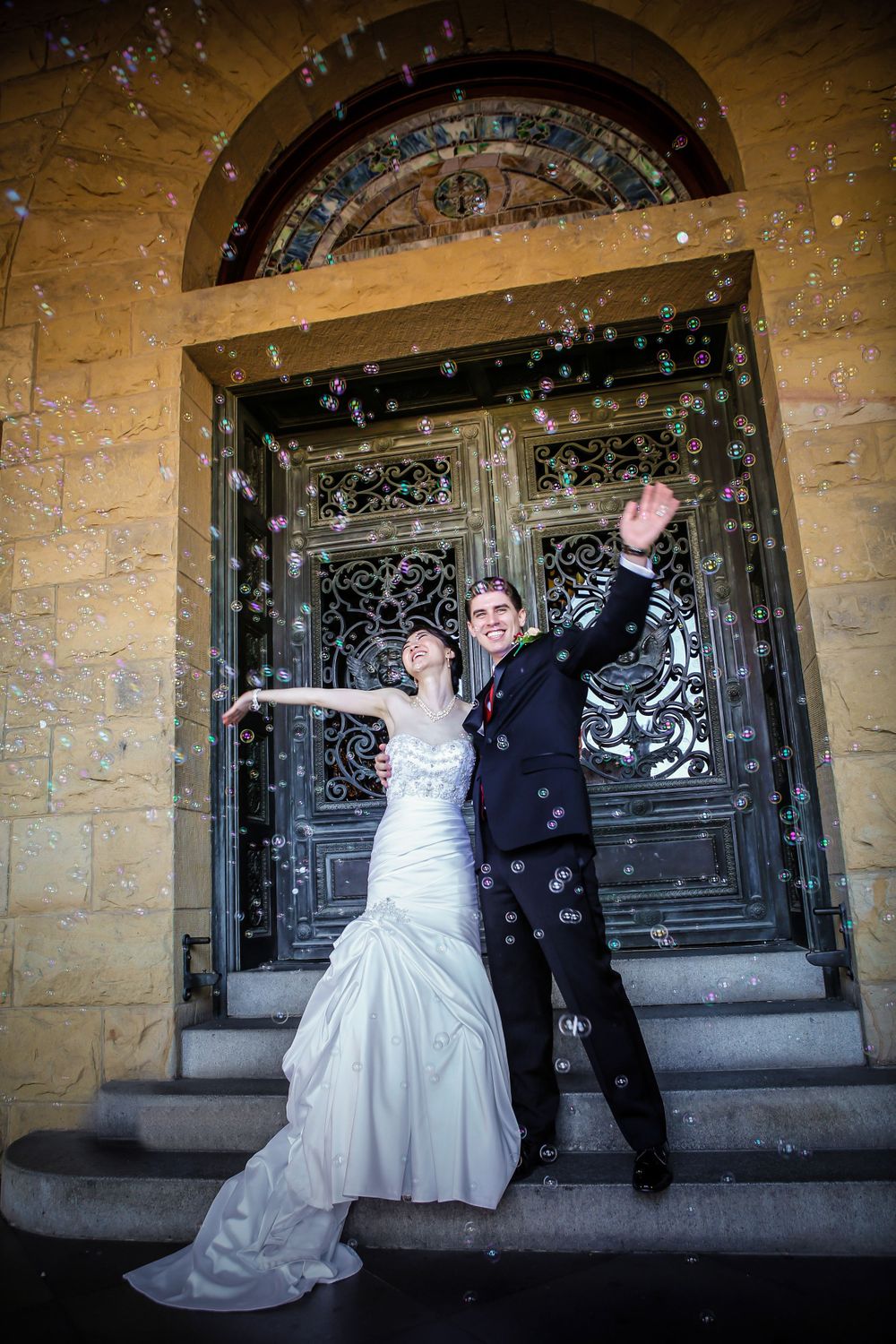 Celebratory wedding couple pose under shower of bubbles at grand doorway entrance.