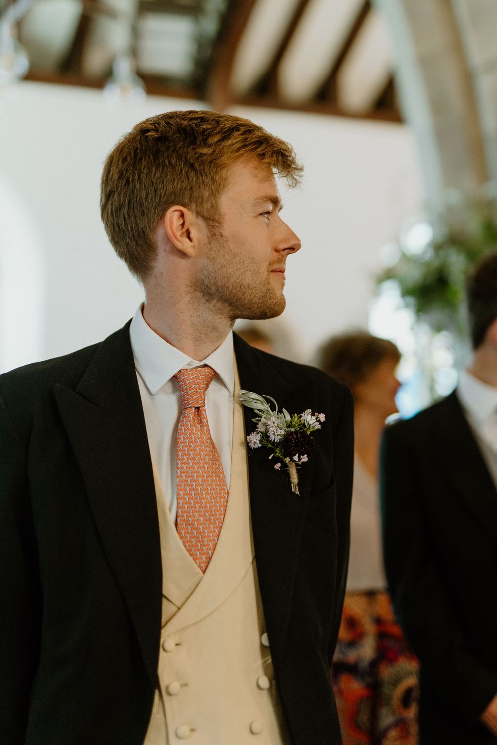 A groom in formal attire with coral tie and cream waistcoat stands during wedding ceremony.