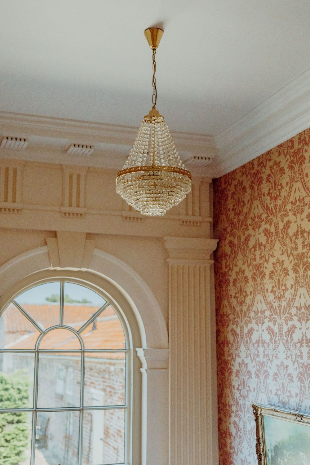 An elegant crystal chandelier hangs from a ceiling in a room with ornate wallpaper and an arched window.