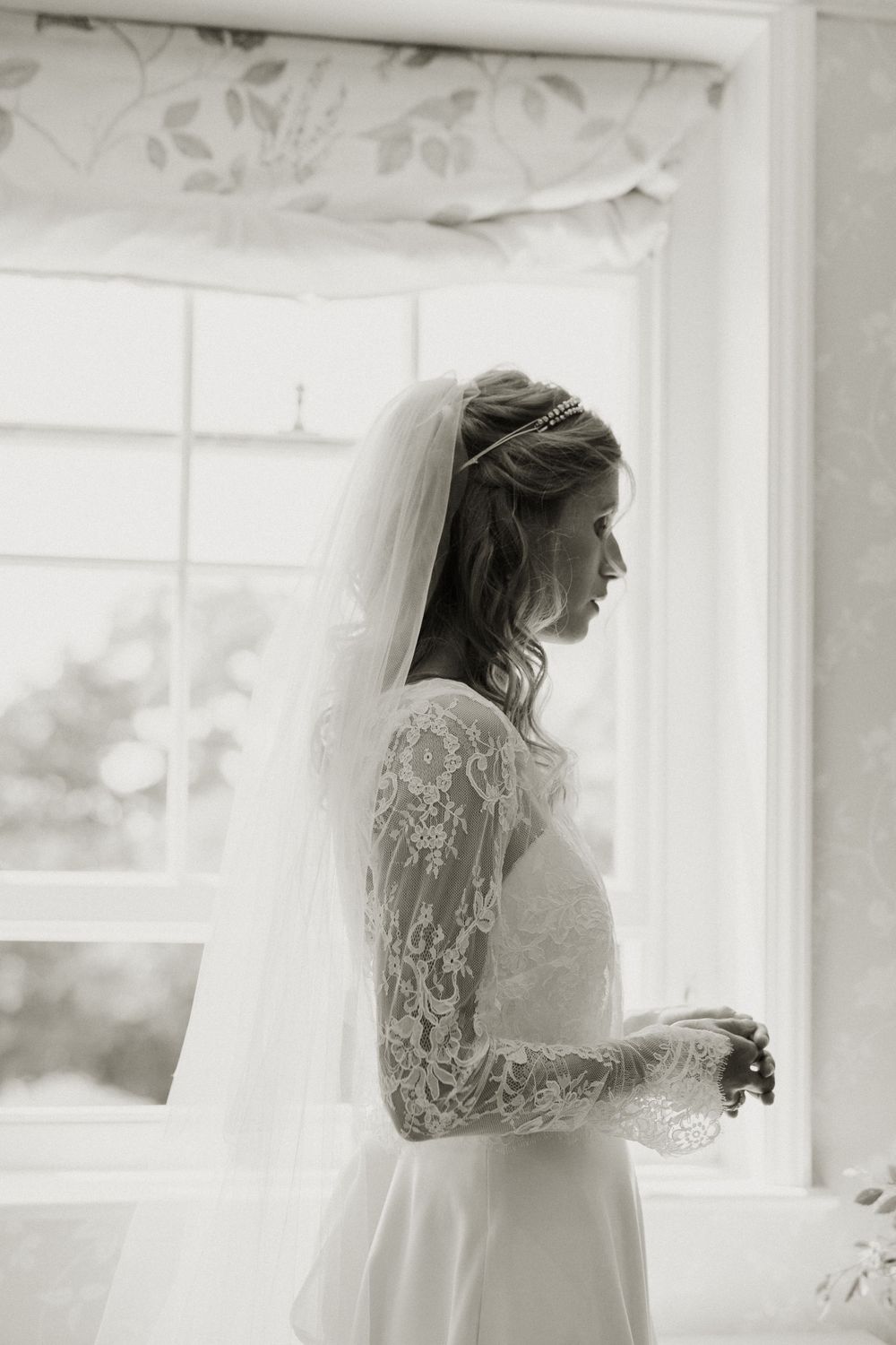 A bride in lace sleeve gown stands in silhouette by window in black and white photograph.