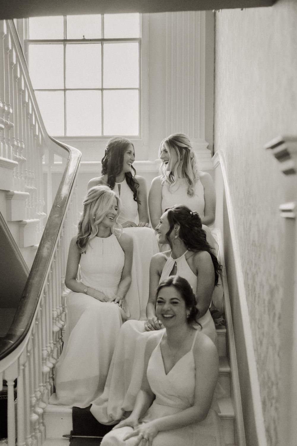 Bridesmaids in light colored dresses sit together on an elegant white staircase in a vintage black and white photo.