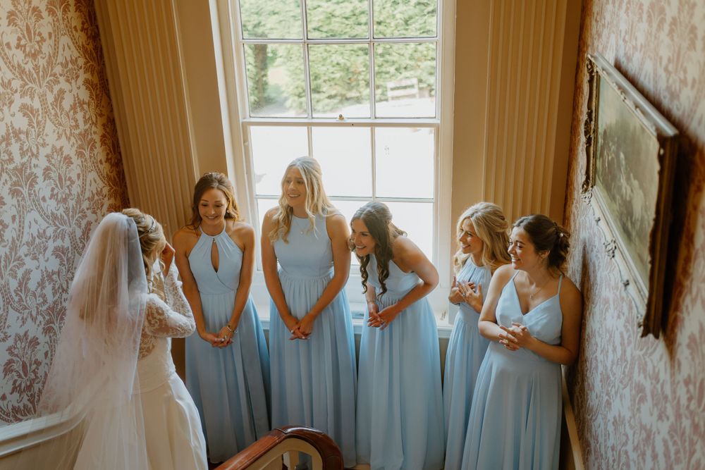 Bridesmaids in pale blue dresses gather around the bride in a sunlit room with patterned wallpaper.