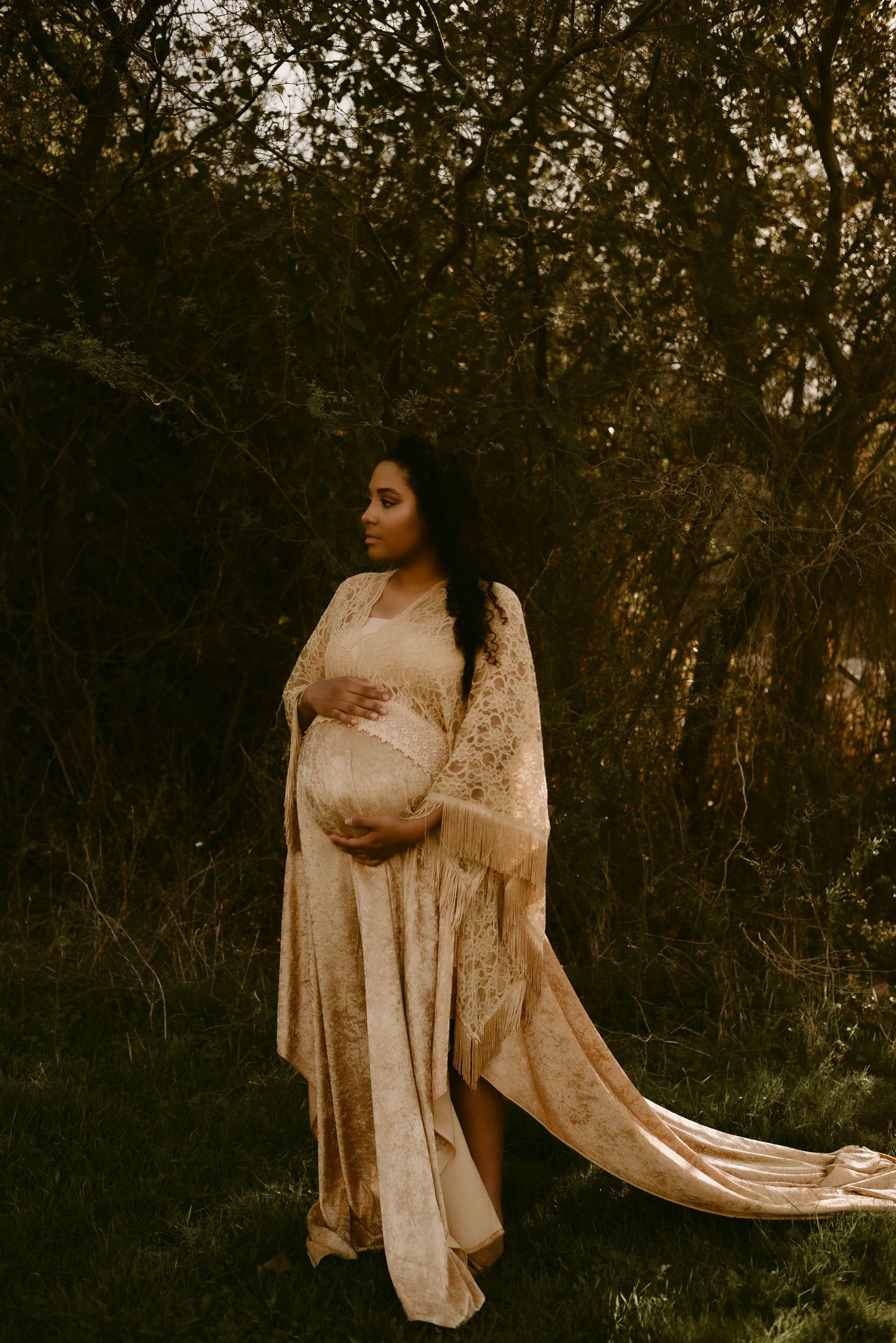 Artistic maternity portrait in flowing cream gown with intricate lace details outdoors.