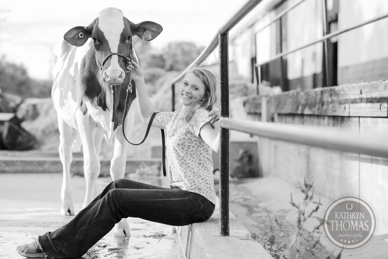 A black and white photo shows a dairy cow and person posing together against a farm railing.