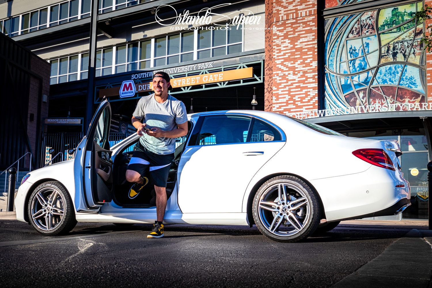 White luxury car parked against brick wall with dramatic lighting and urban backdrop.