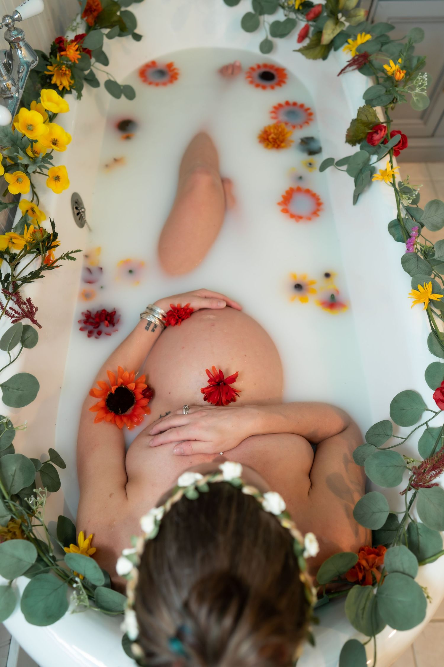 Artistic milk bath photography with colorful flowers and eucalyptus leaves floating in white water.
