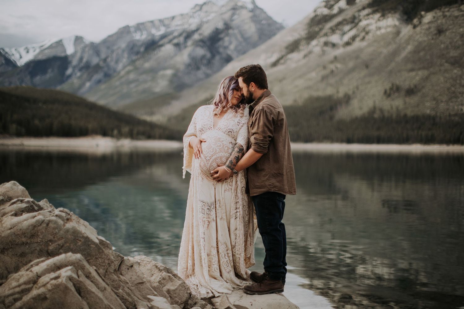 A romantic maternity photo by a pristine mountain lake with snow-capped peaks in the background.