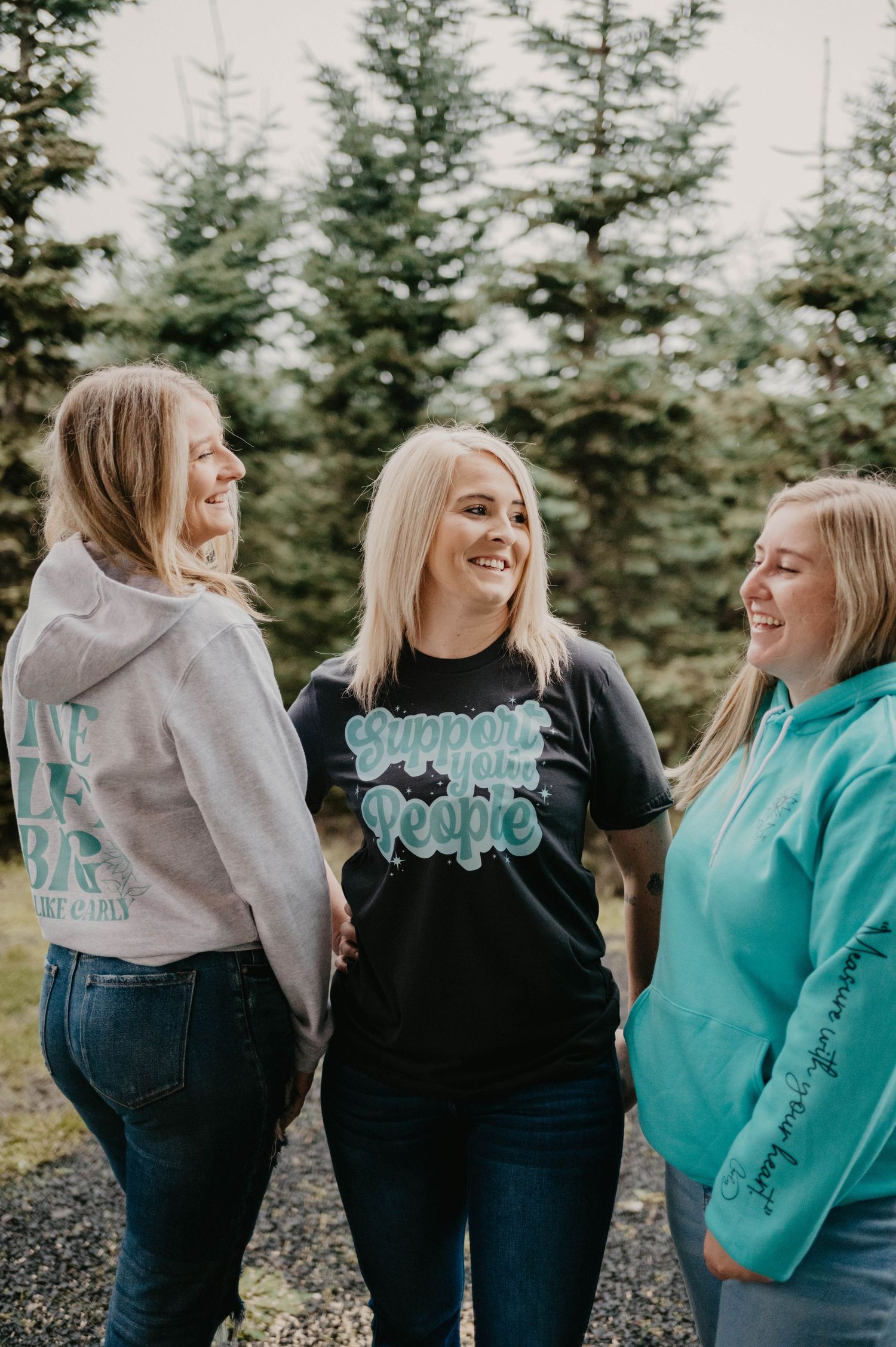 Three friends laughing together outdoors with evergreen trees in background.