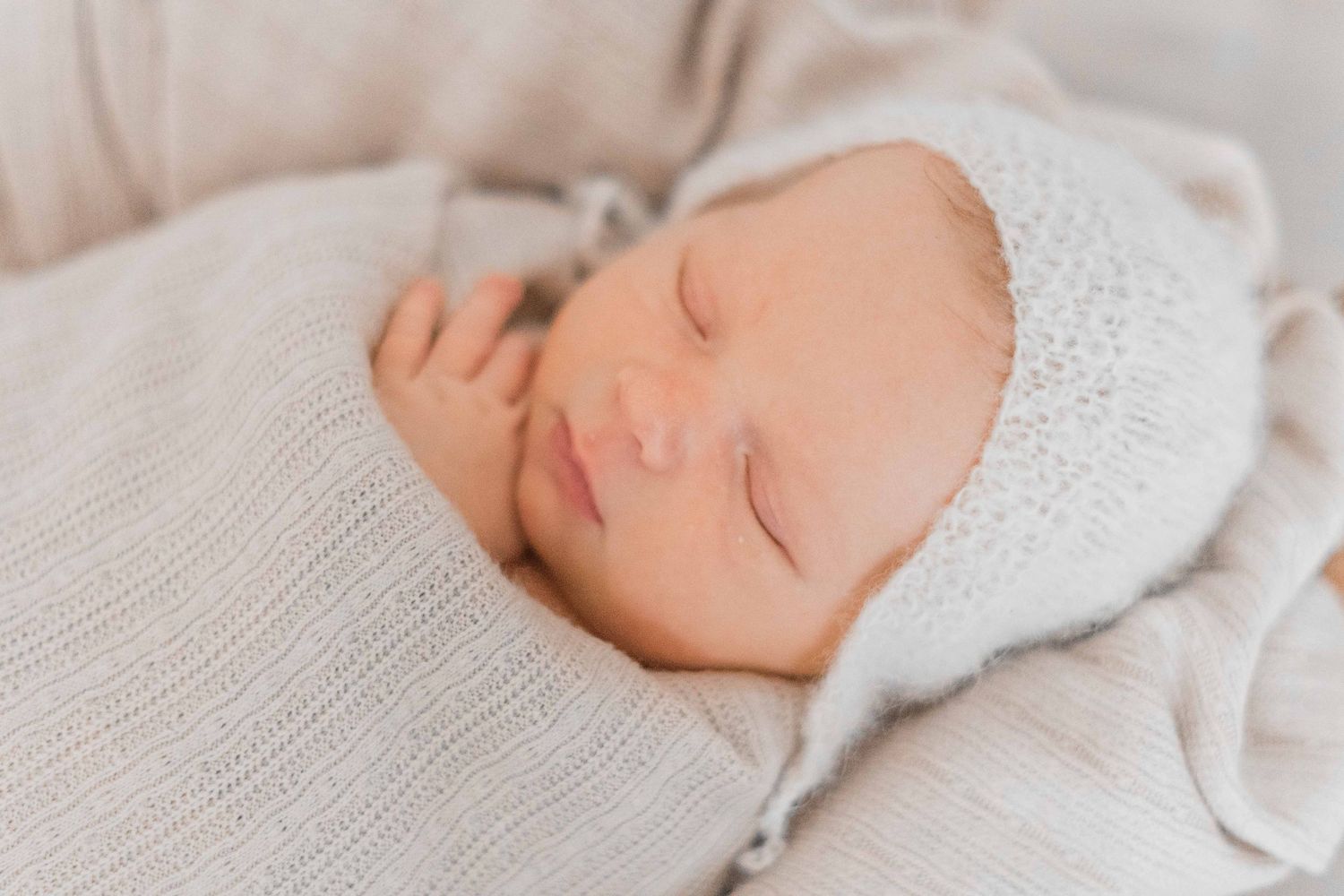 Sleeping newborn baby wearing white knit bonnet and swaddle blanket.