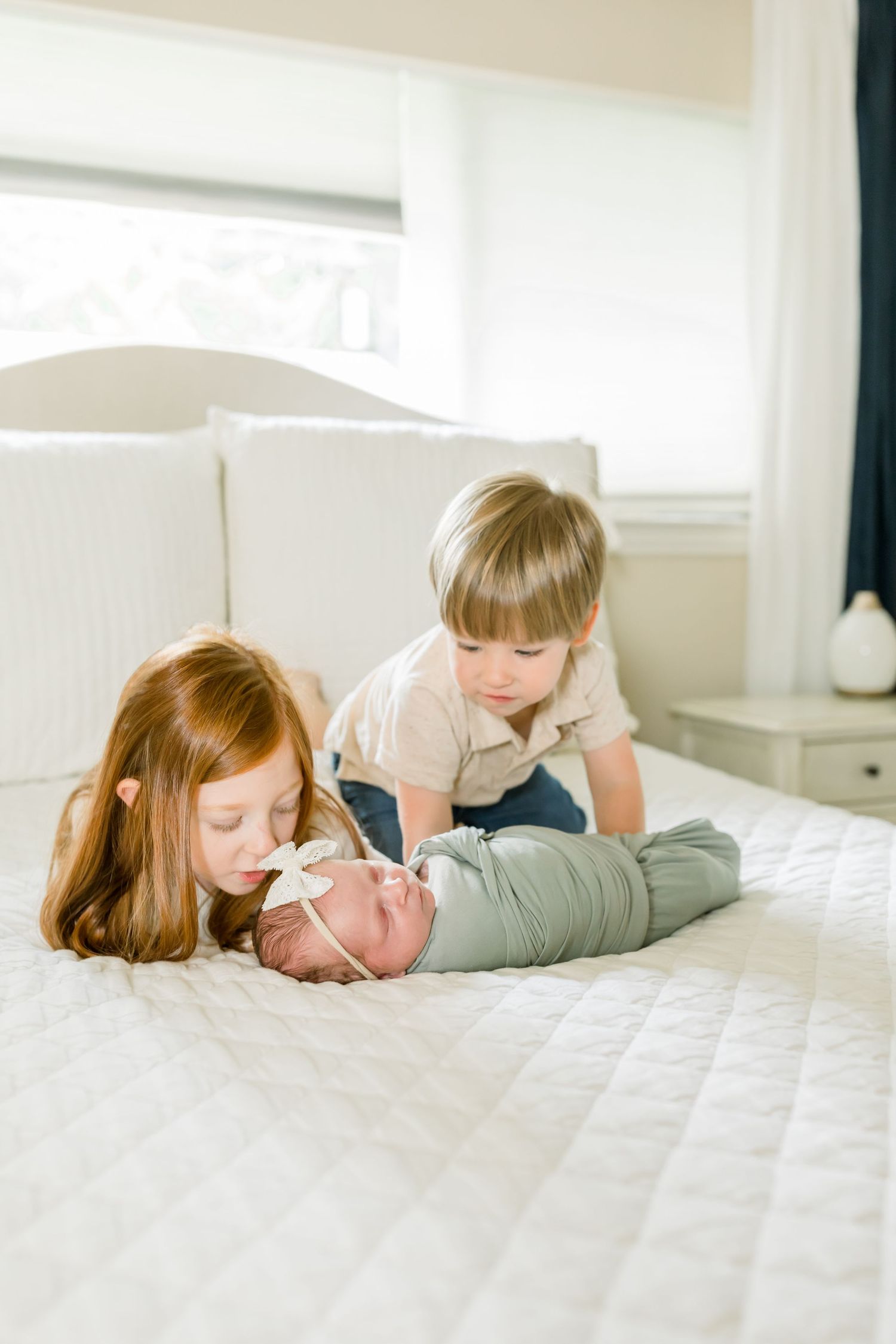 Two young siblings gently play with their swaddled baby sibling on a white bed.