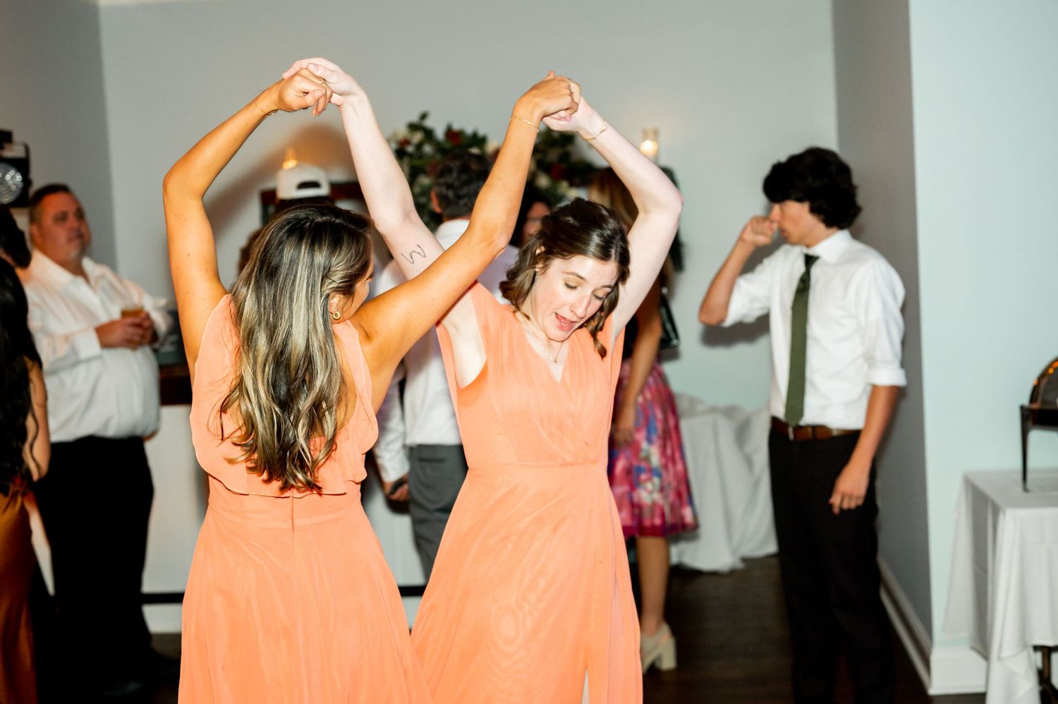 Bridesmaids in coral colored dresses dance together at a wedding reception.