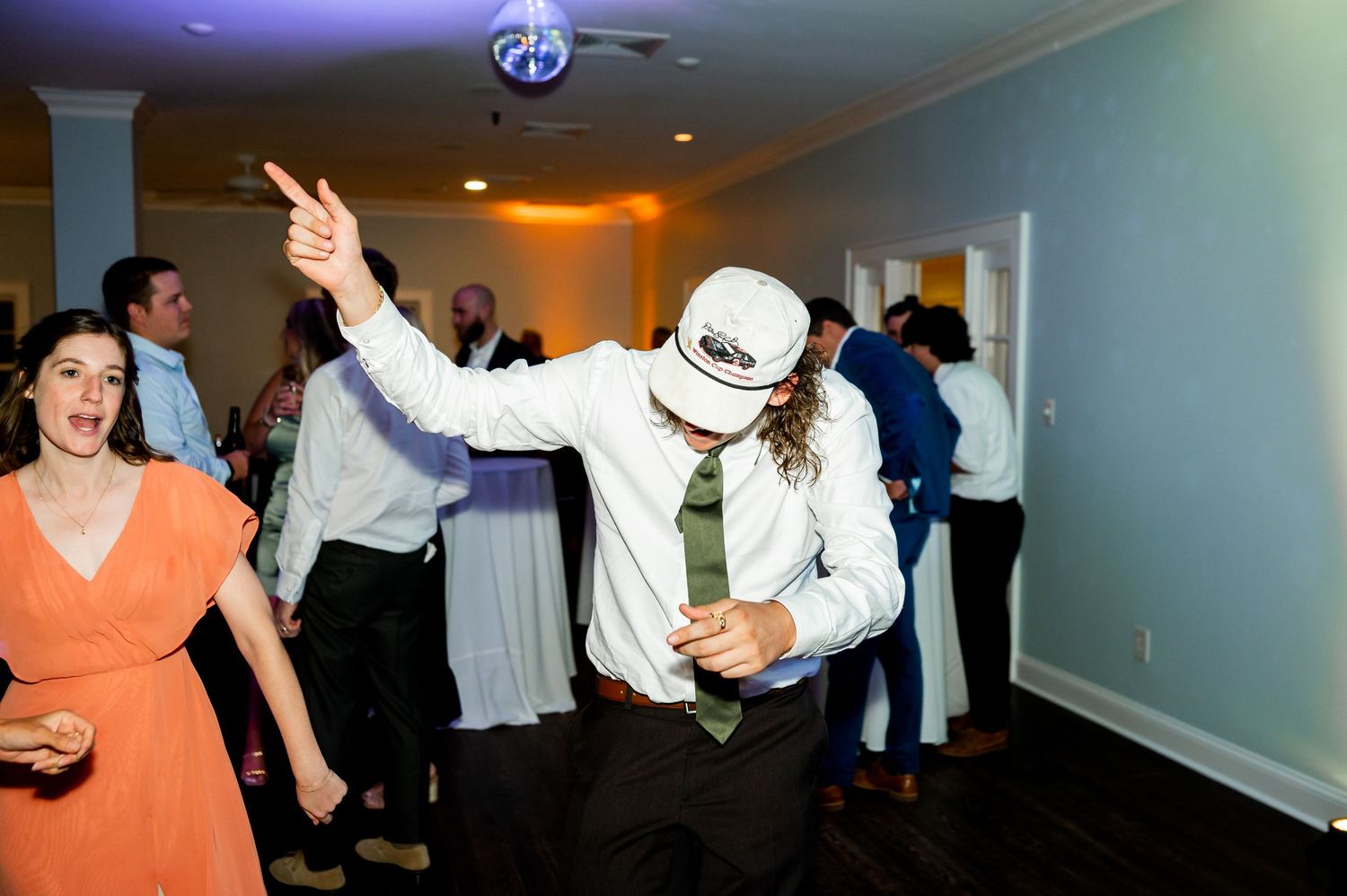 Wedding guests dancing energetically at a reception in a white-walled venue.