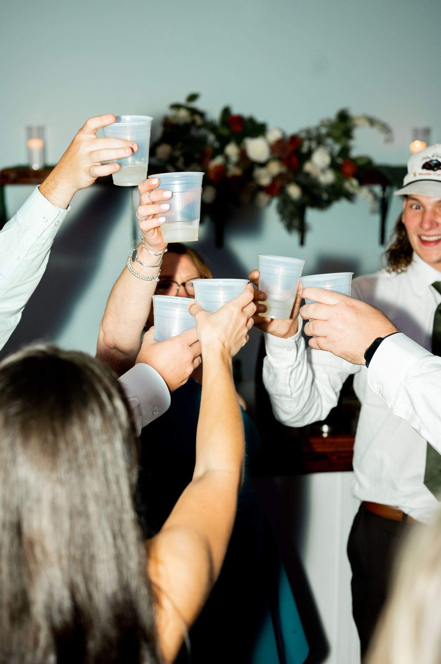 Wedding guests raise plastic cups in a celebratory toast with floral decorations in background.