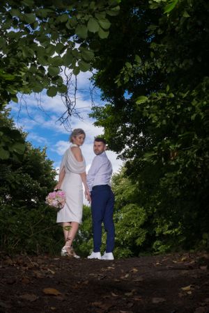 A couple poses on a forest path with blue sky peeking through the trees.