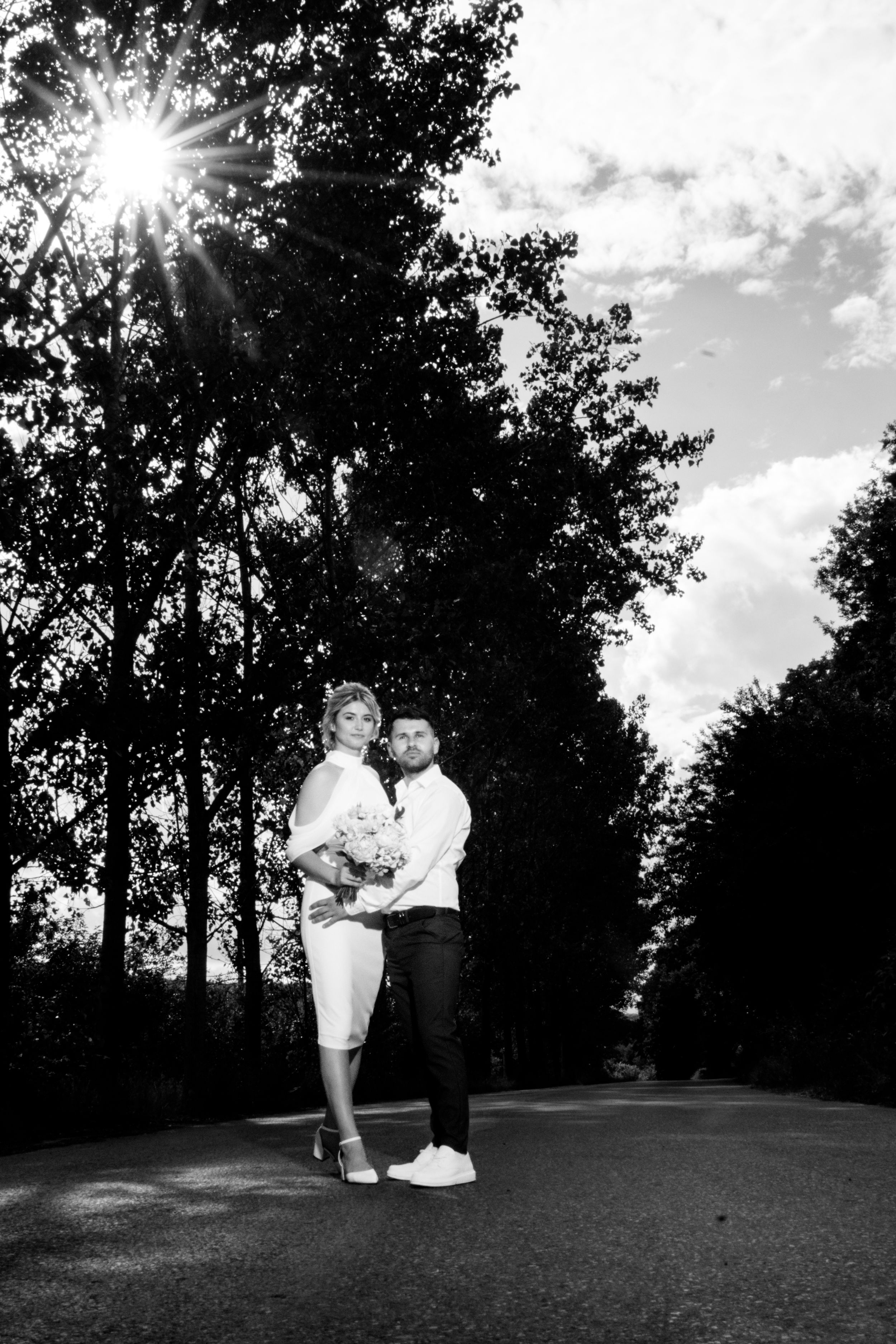 Black and white portrait of couple standing on path with sunburst through trees.