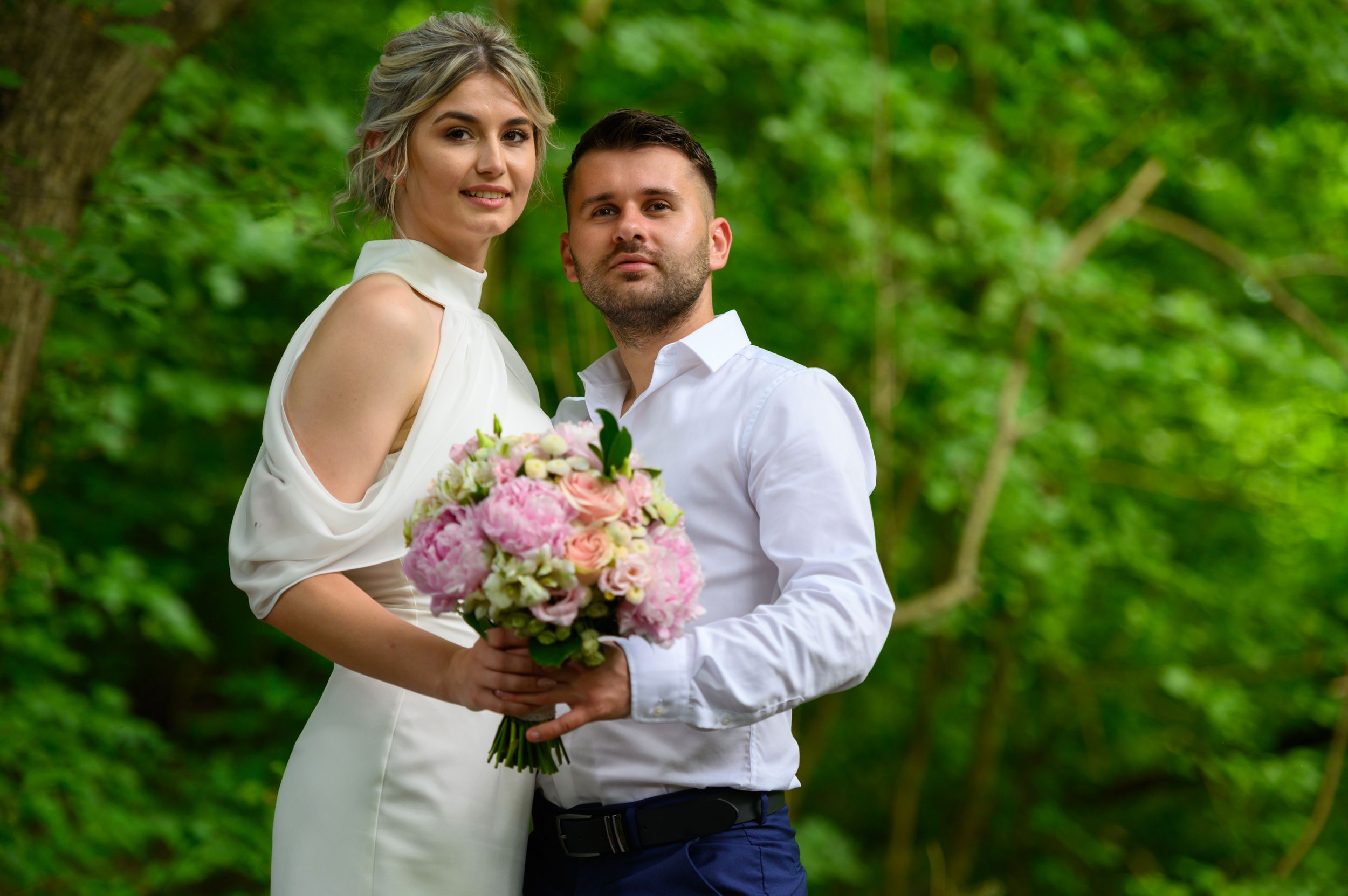 Couple in wedding attire pose with pink bouquet in lush green garden setting.