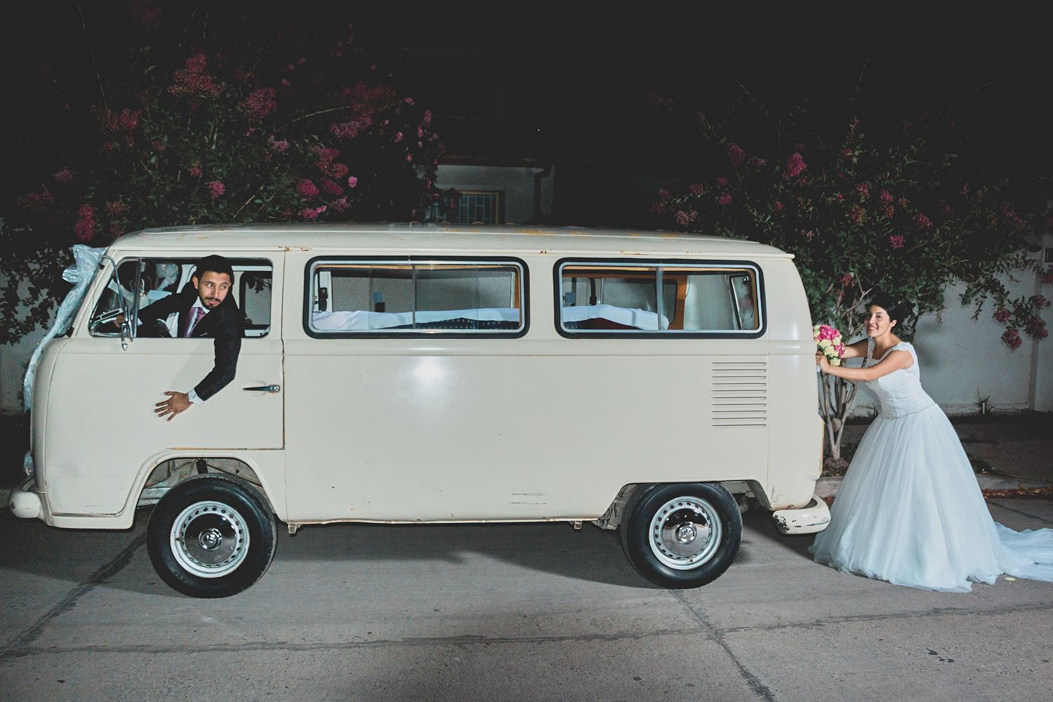 Wedding couple poses with classic cream-colored Volkswagen van in urban setting.