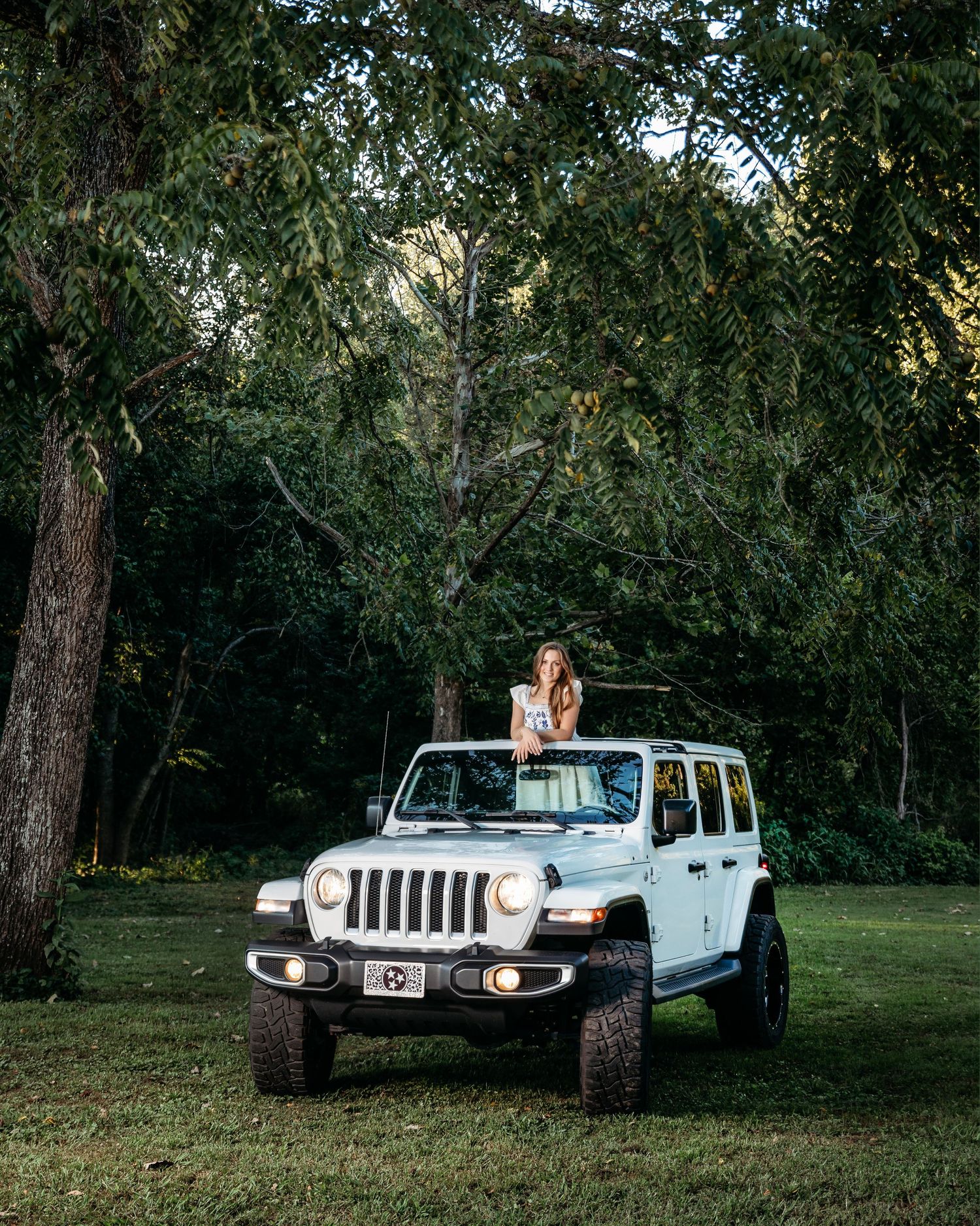 Senior girl in white Jeep parked among trees in a natural outdoor setting at dusk. Middle TN senior photographer