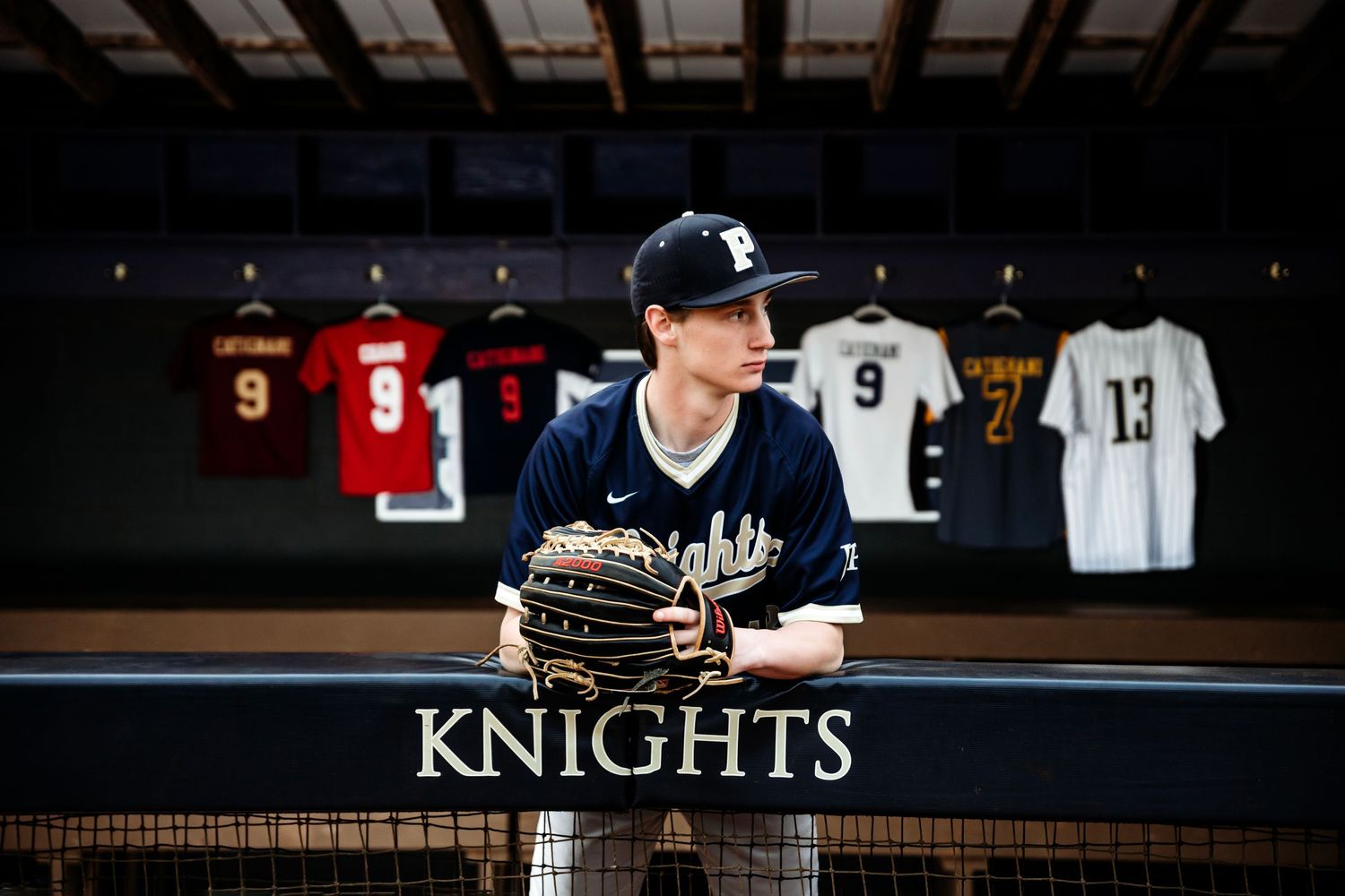 Baseball player stands in dramatic lighting with team jerseys displayed in the background. Middle TN senior photographer
