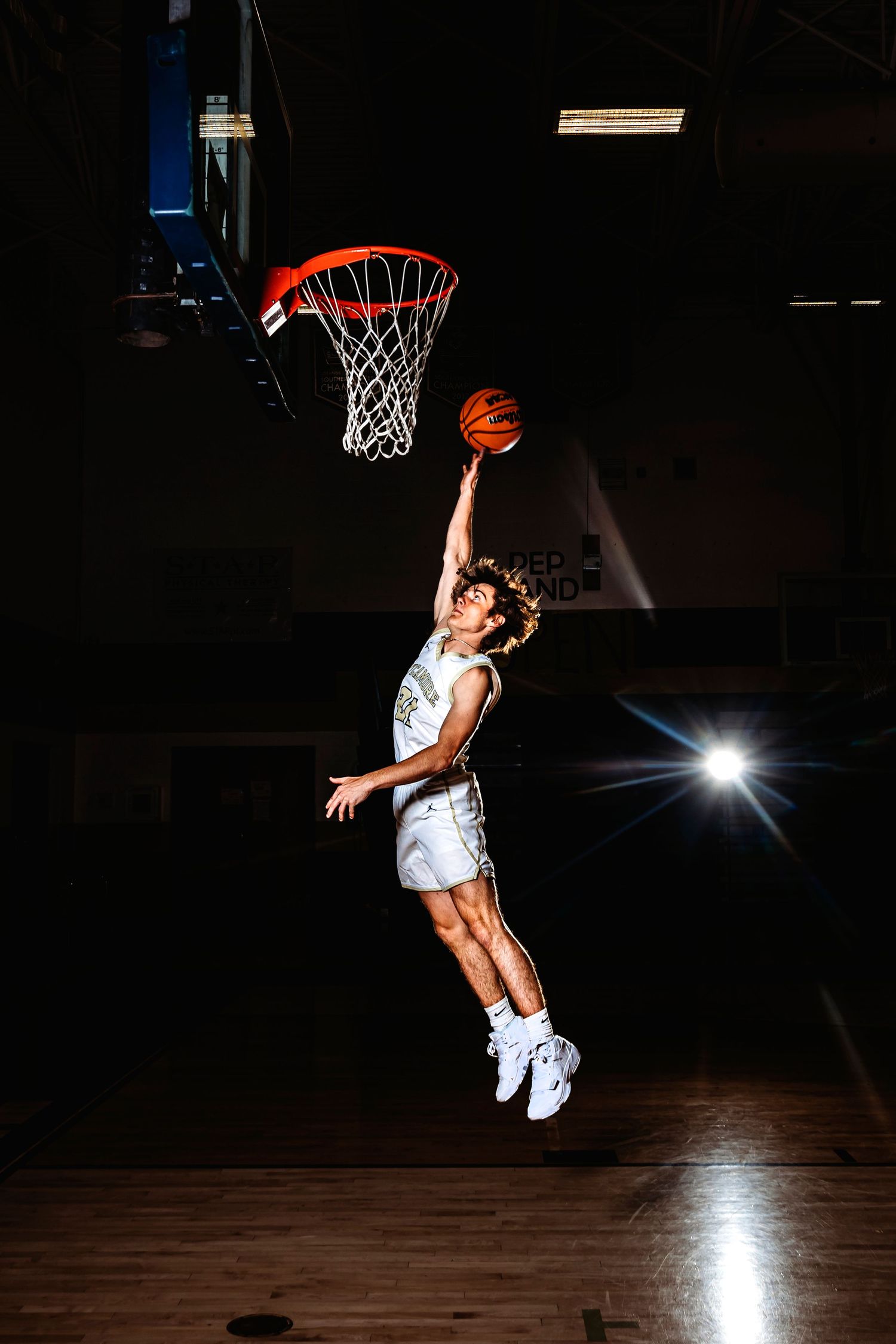 Basketball player performs an athletic slam dunk in a dark gymnasium with dramatic lighting. Middle TN senior photographer