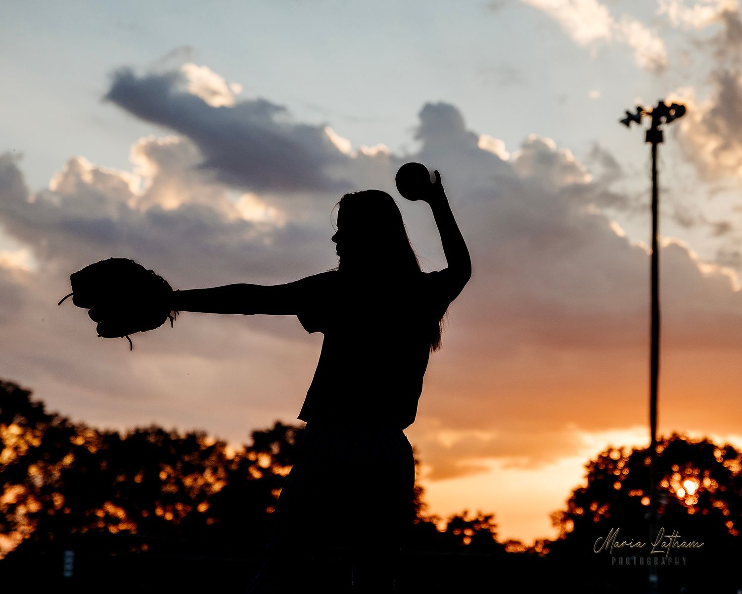 Silhouette of a baseball pitcher in mid-throw against a dramatic sunset sky with stadium lights. Middle TN senior photographer