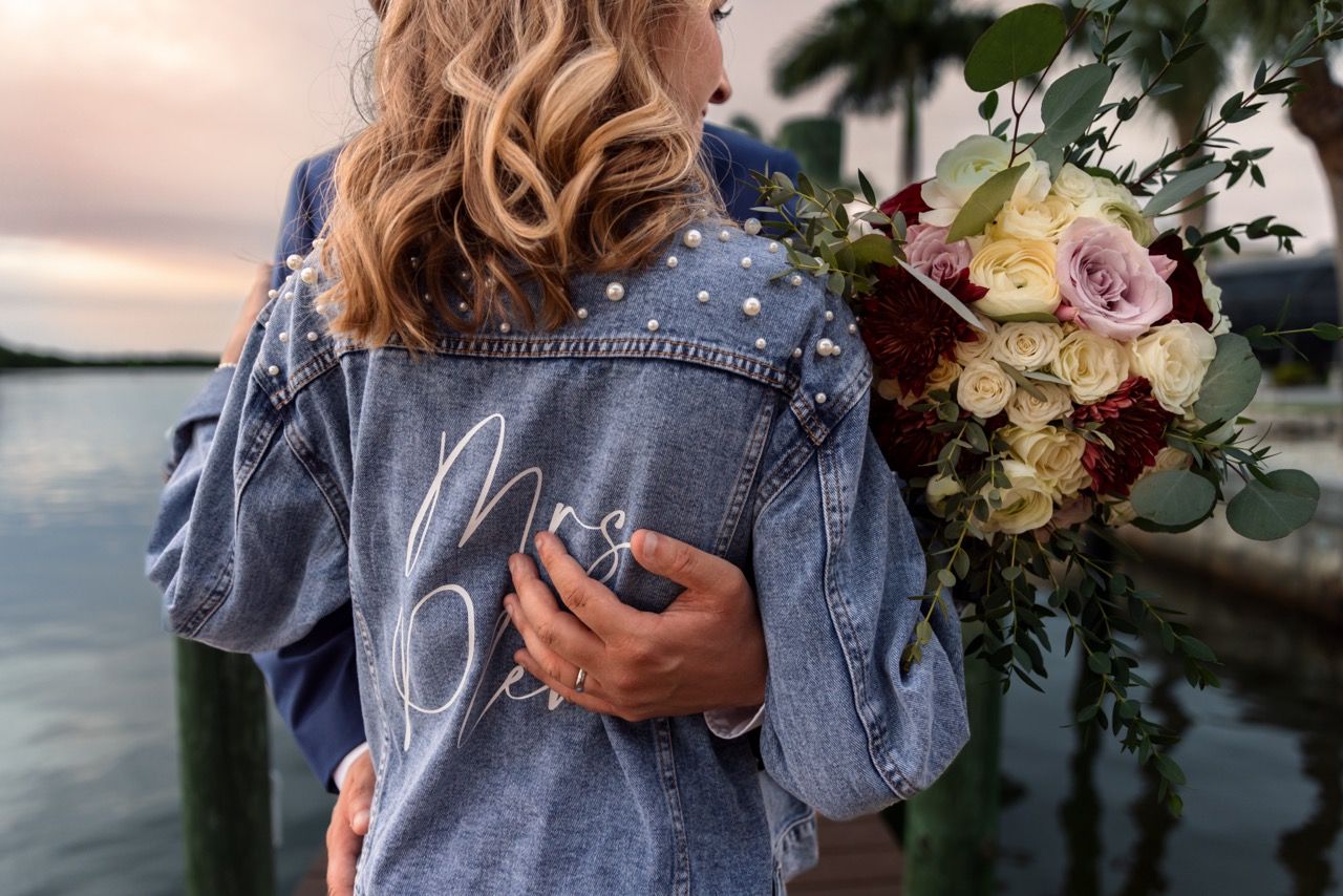 Denim jacket with decorative detail holds flowers against sunset waterfront backdrop.