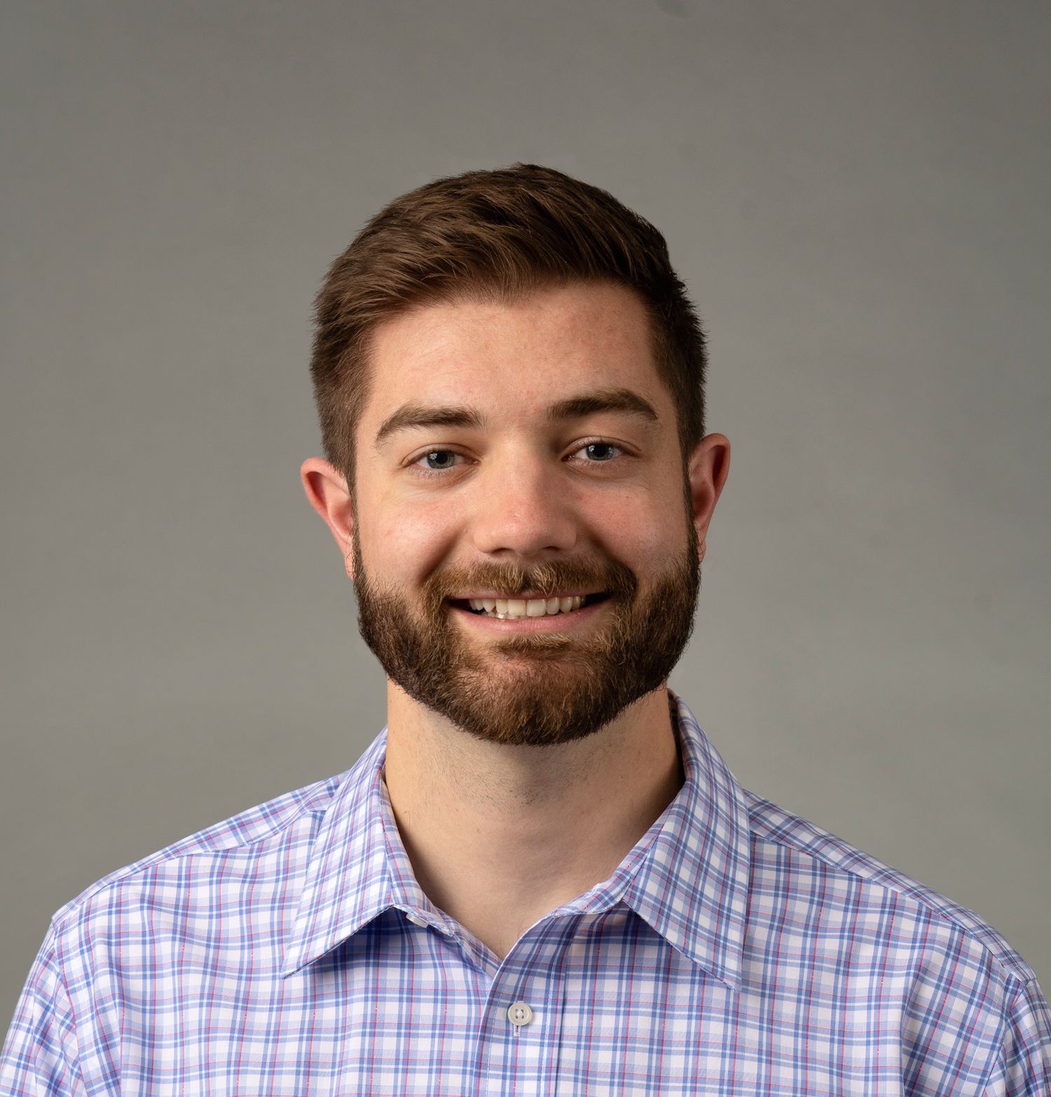 Professional headshot of person wearing a light blue checkered dress shirt against a gray background.