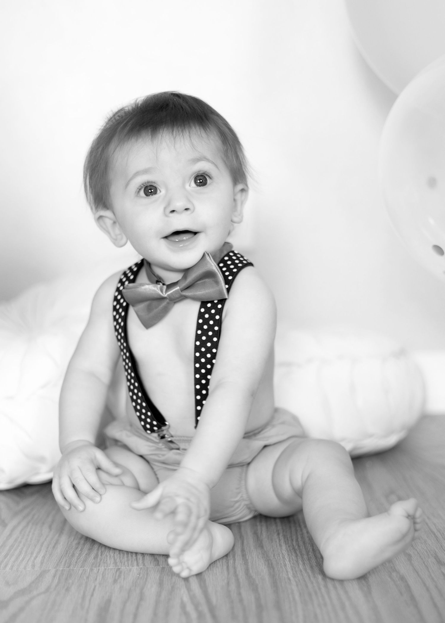 Baby in polka dot suspenders and bow tie sitting on floor with white backdrop.