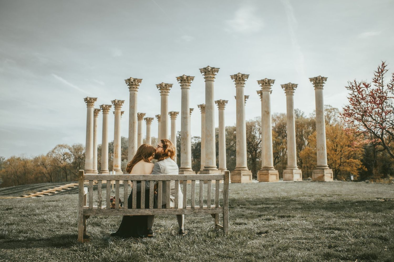 Ancient Roman columns stand majestically in a field with bench in foreground during cloudy sunset.