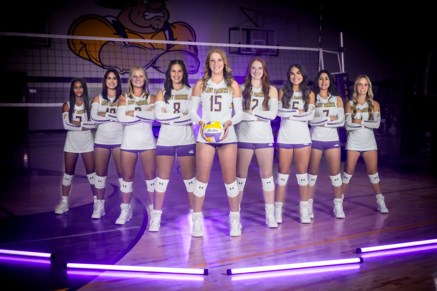 High school volleyball team poses for team photo on purple-lit court with team mascot logo visible in background.