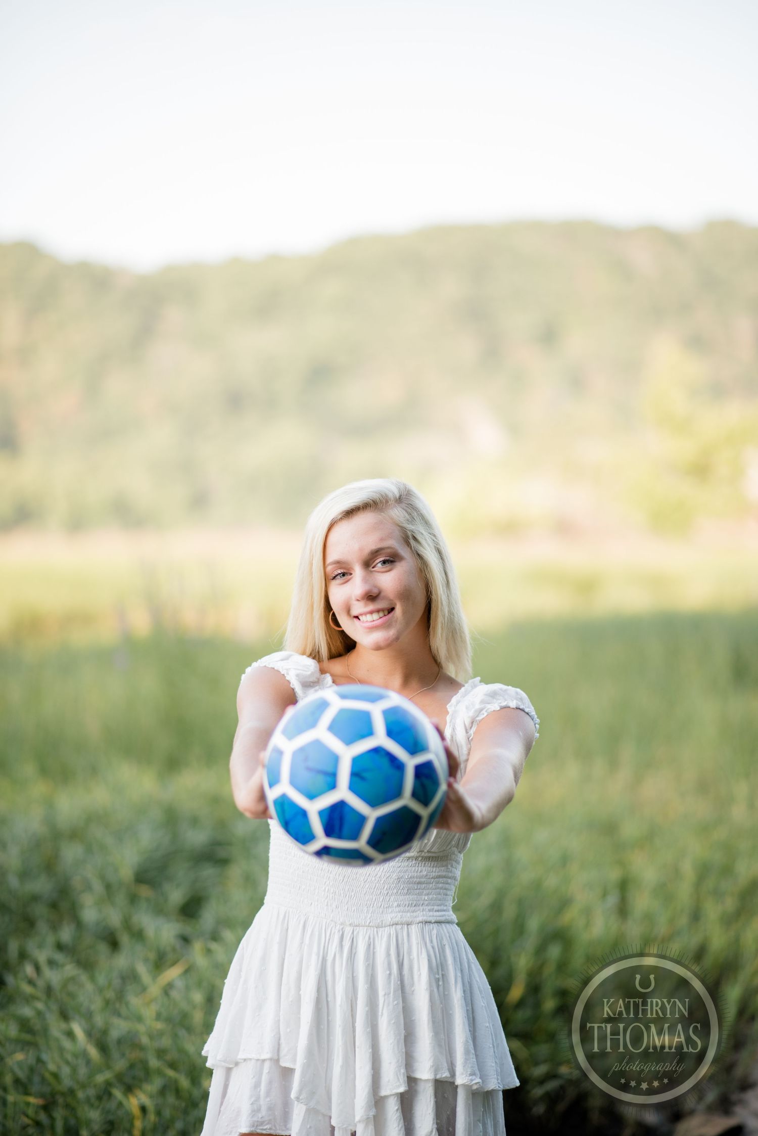 Woman in white dress holds up a blue and white soccer ball against a mountain backdrop.