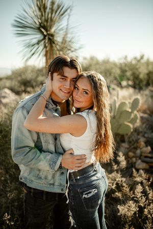 A happy couple embraces against a desert landscape with cacti and yucca plants behind them.