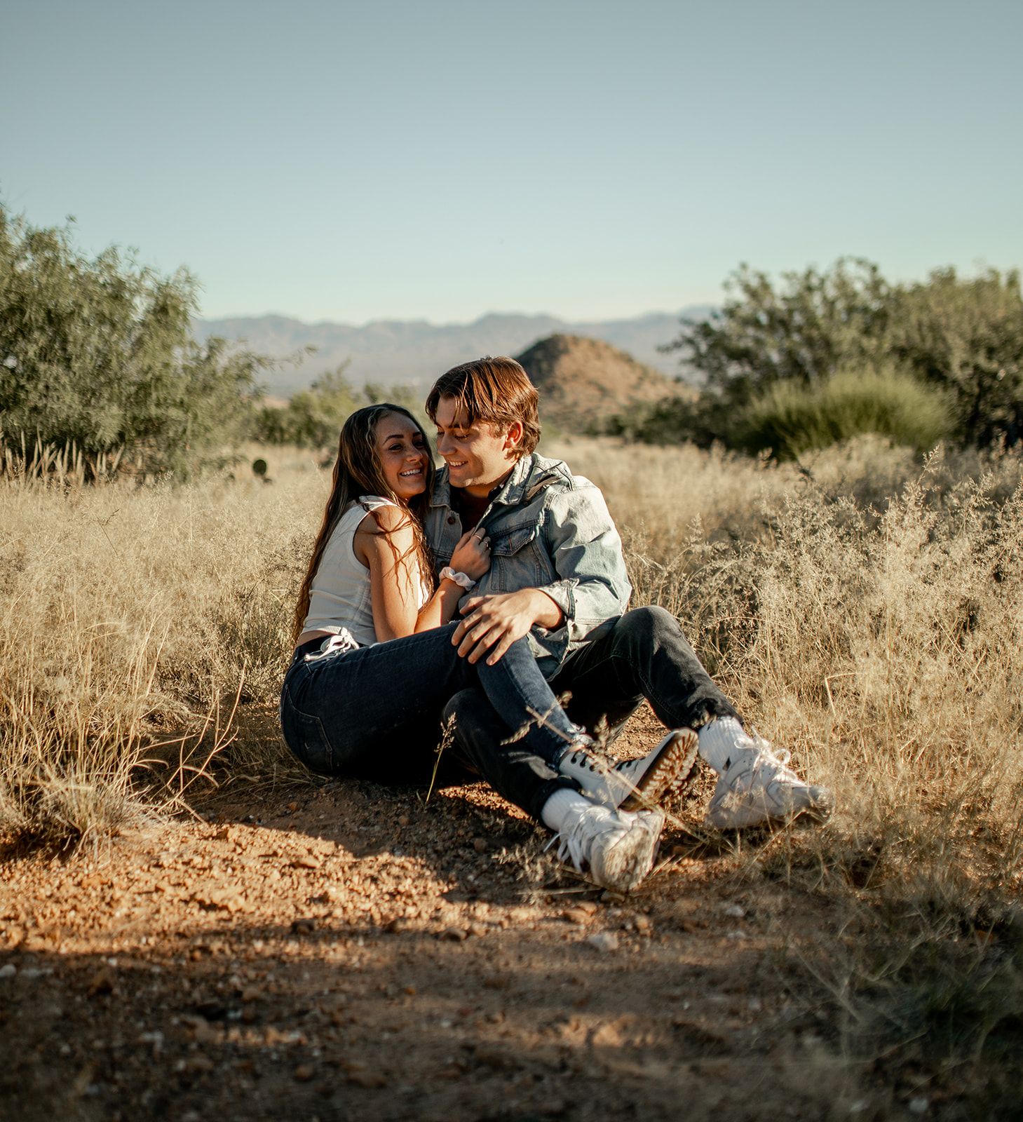 A couple rests together on a desert trail surrounded by dry grass and mountains.