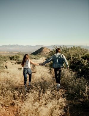 A couple walks hand in hand through desert brush with mountains in the distance.
