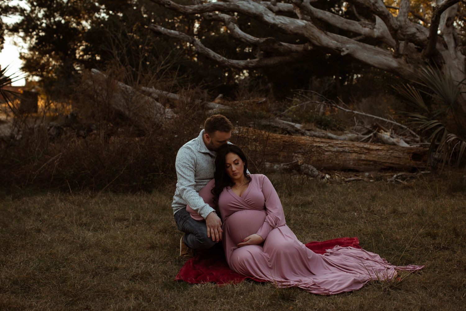 A romantic maternity photoshoot at sunset with a couple sitting on a red blanket under a twisted tree.