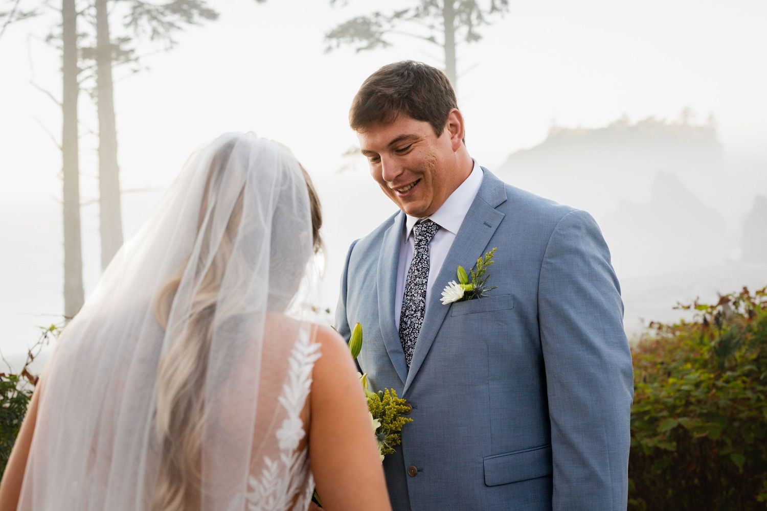 Smiling groom in blue suit shares first look moment with veiled bride on foggy Pacific Northwest coastline.