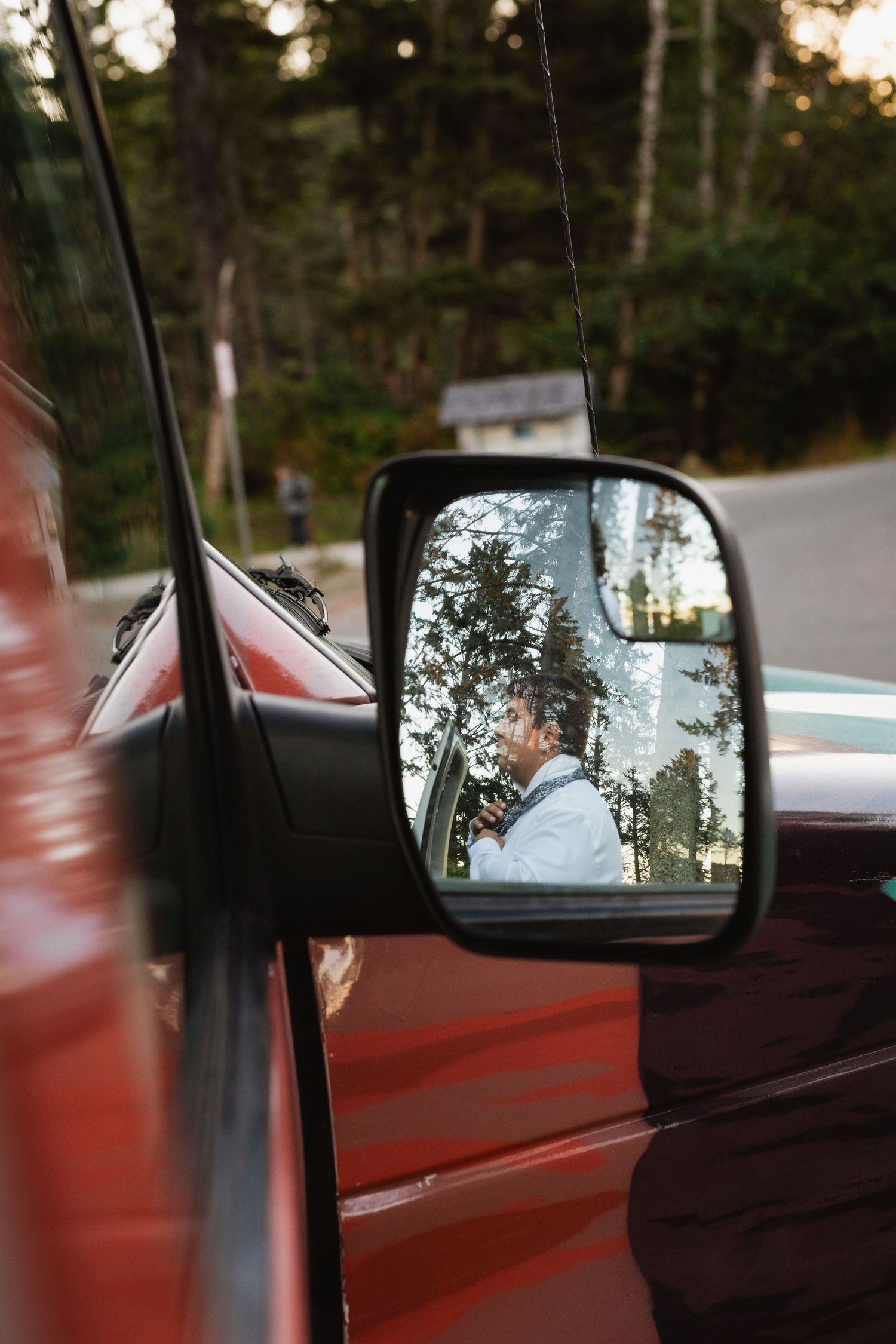 Bride in wedding dress reflected in a red truck side mirror surrounded by forest trees.